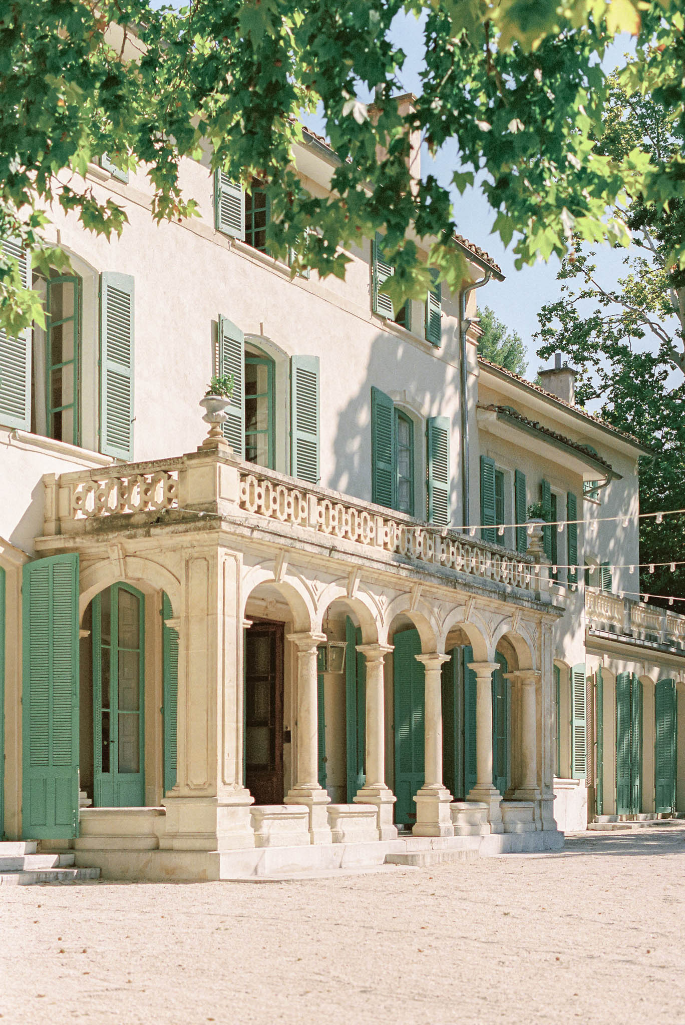 French bastide manor house with sage green shutters, stone arched colonnade, and gravel courtyard