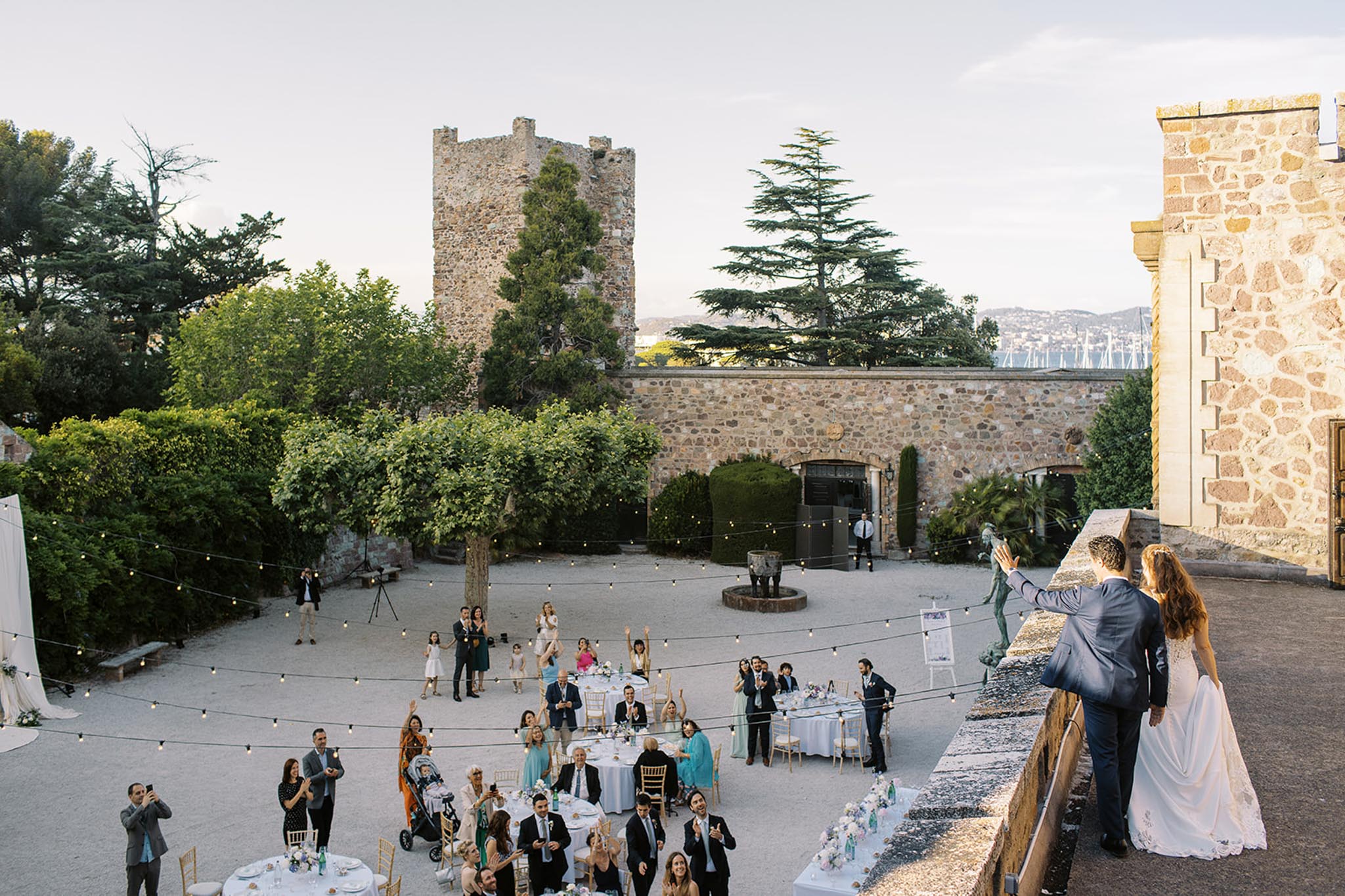 Bride and groom entering courtyard reception with festoon lights gold chairs and medieval tower at dusk