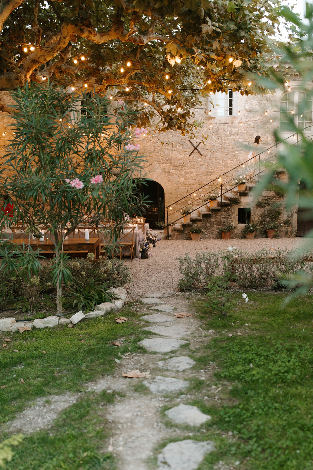Dusk reception setup in Provencal stone courtyard with wooden tables, rattan chairs, and bistro string lights