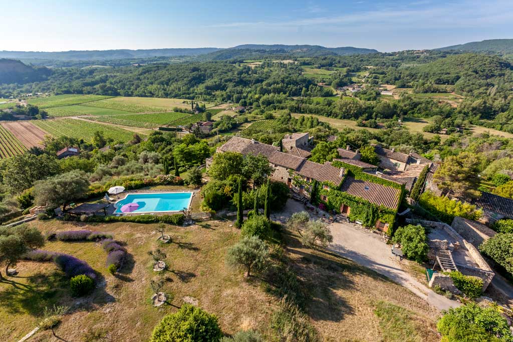 Aerial view of stone farmhouse complex with pool, lavender, and vineyard rows in rolling landscape