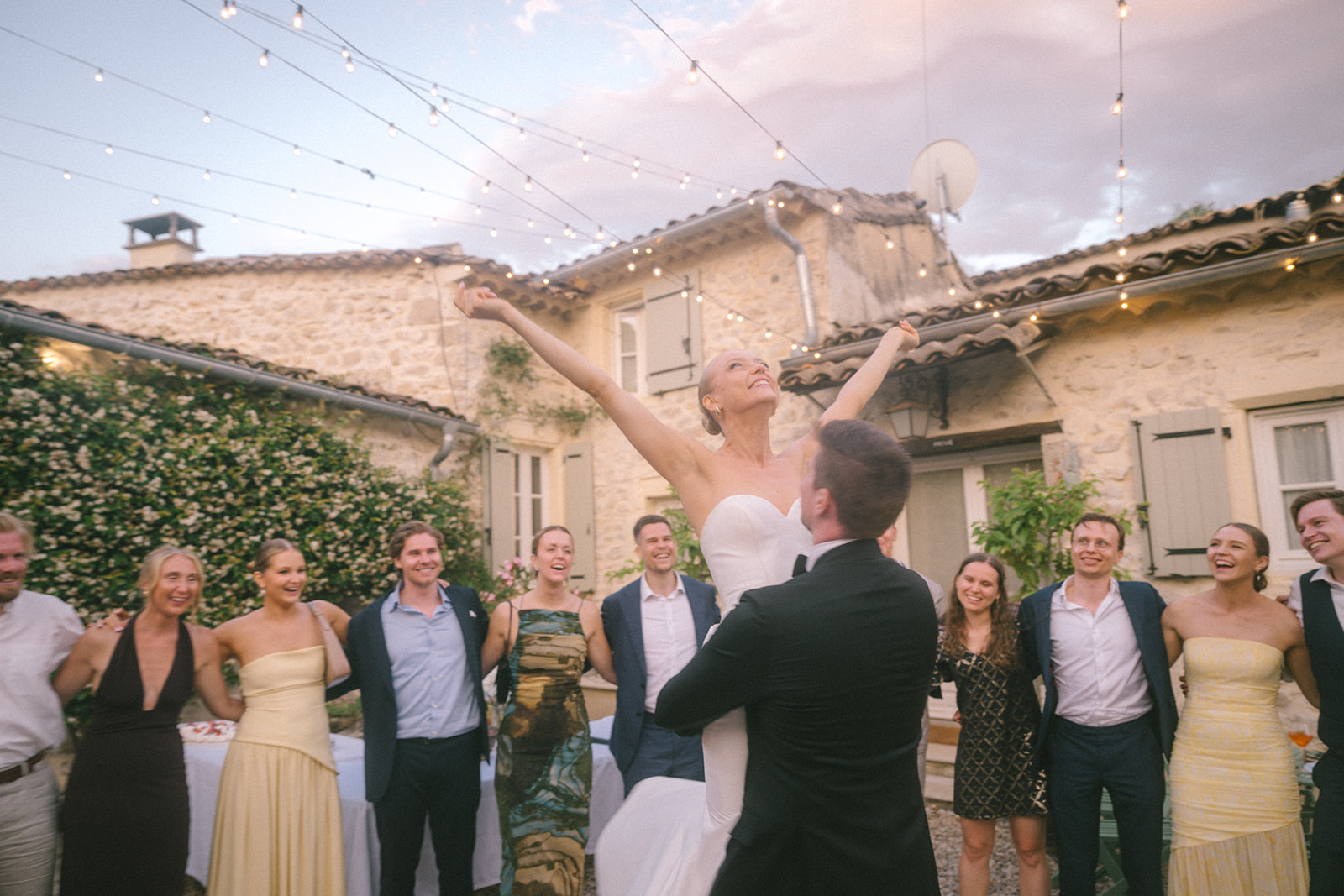 Groom lifts bride during first dance as ten guests circle them under bistro lights in Provencal courtyard