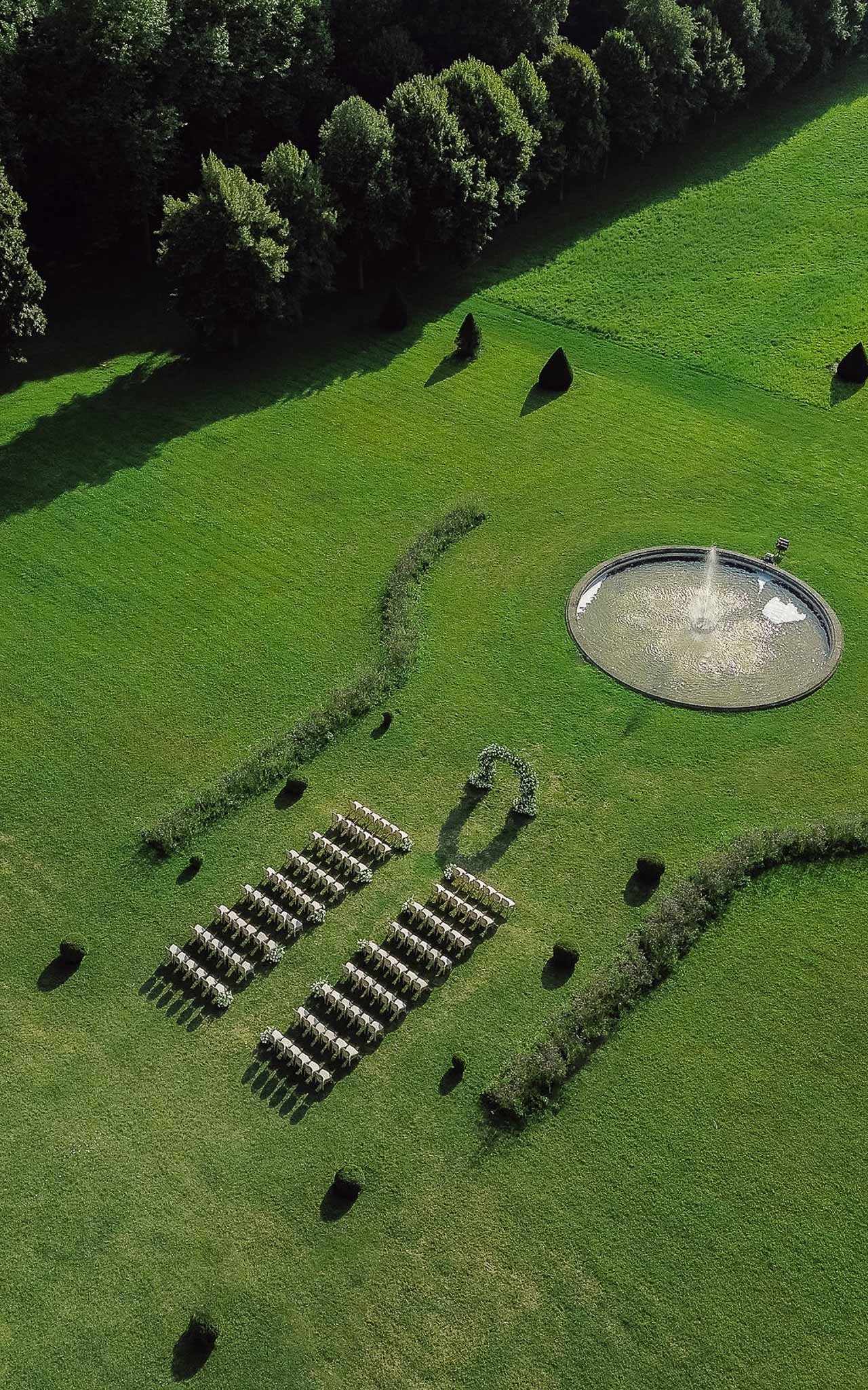 Aerial ceremony setup with white chairs facing floral arch beside circular fountain in formal garden