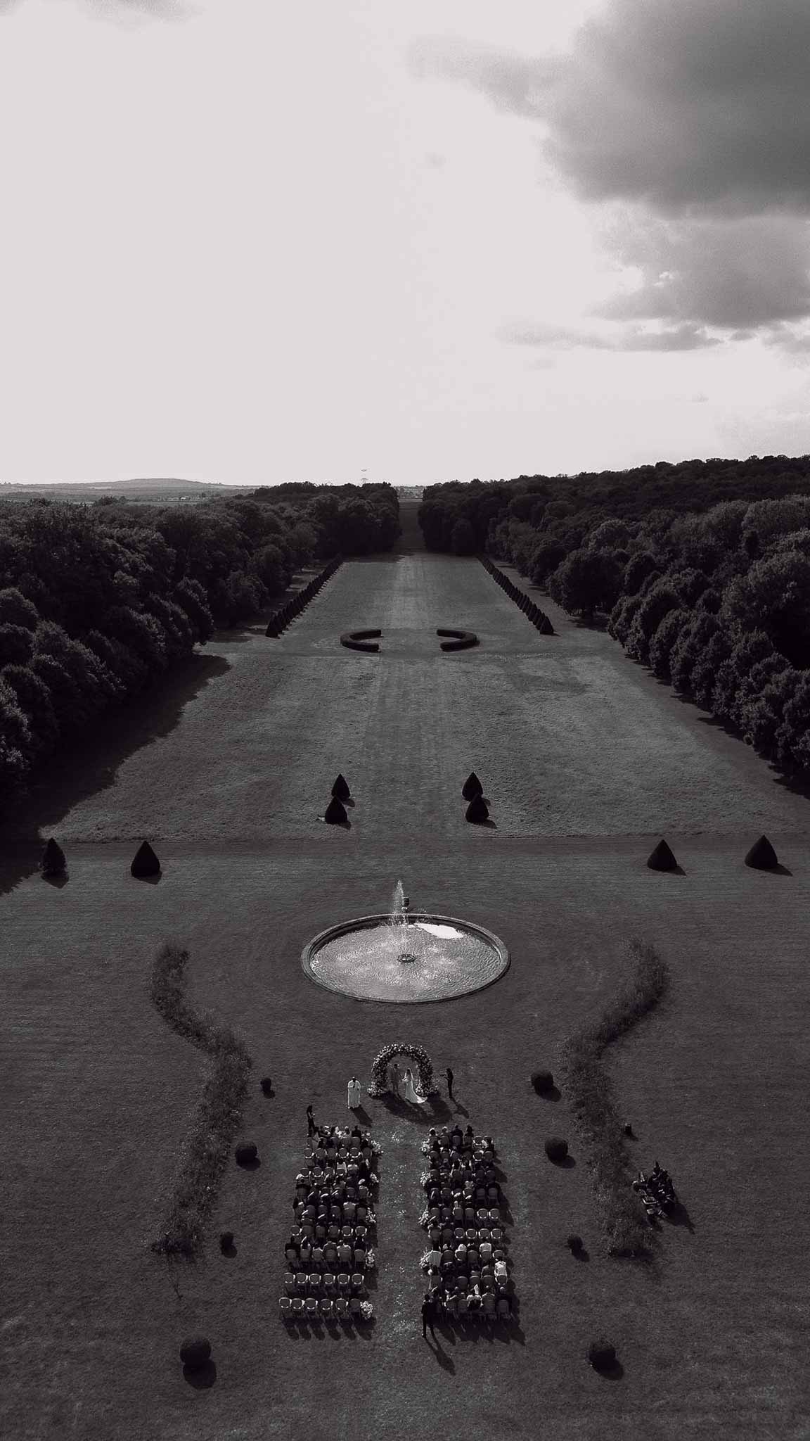 Black and white aerial of 150 guests at ceremony with floral arch beside fountain in formal topiary garden