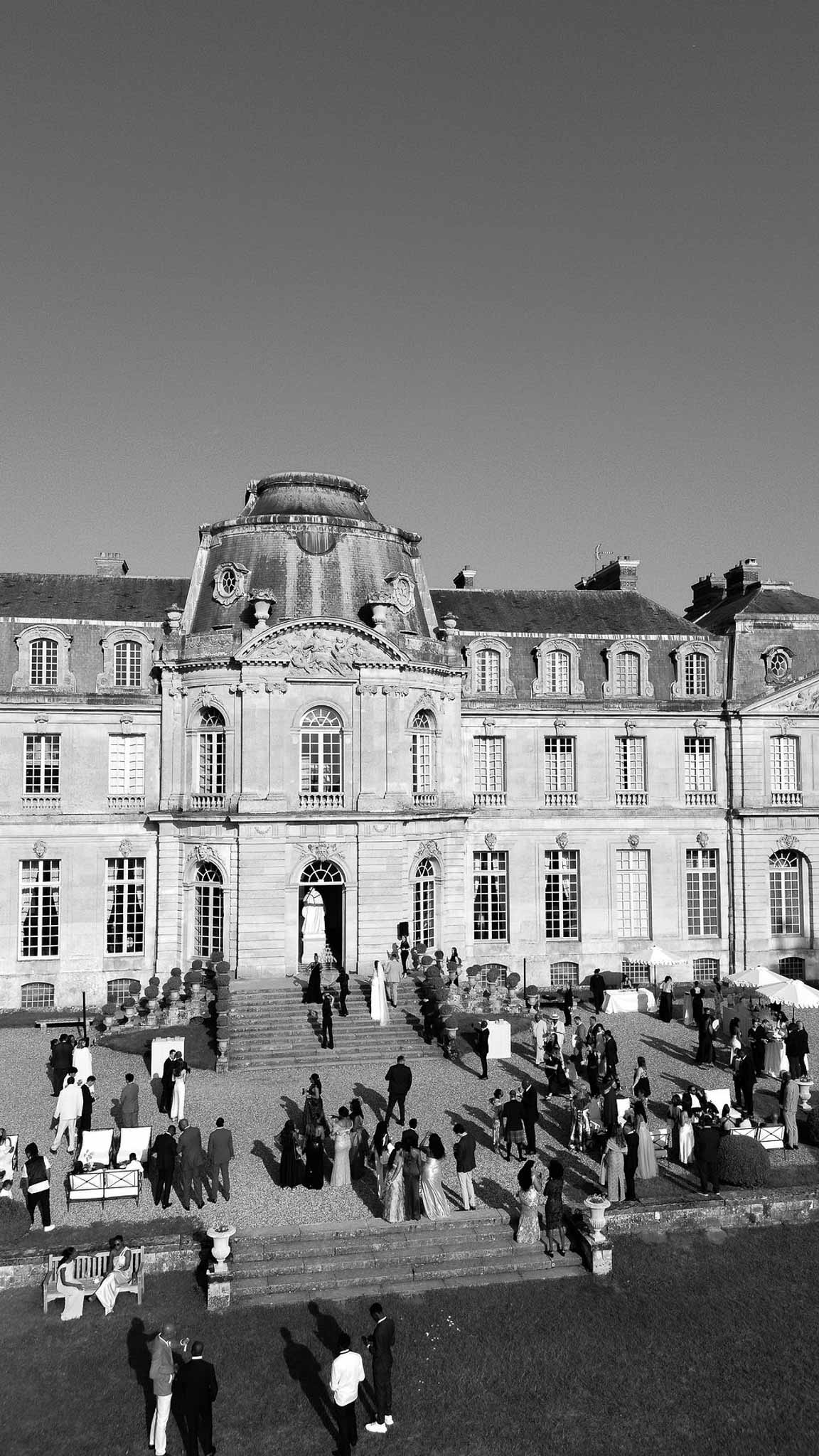 Black and white aerial view of 70 guests at cocktail hour on grand chateau forecourt with mansard dome