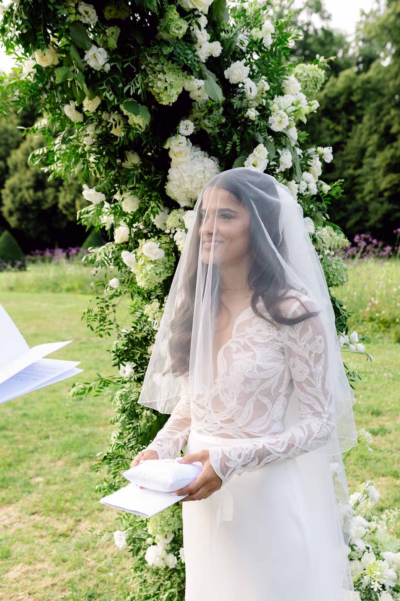 Bride in long-sleeve lace gown and veil reading vows before circular white hydrangea and rose arch
