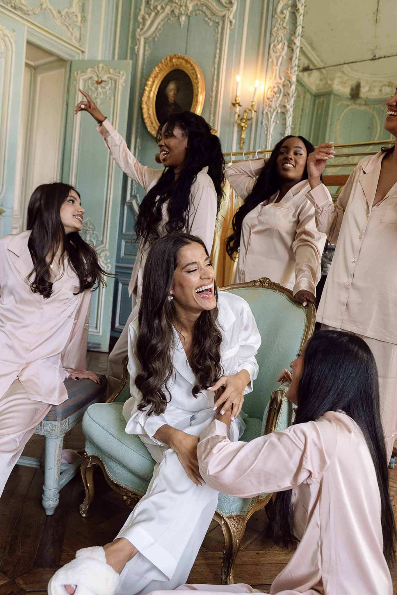 Bride in white pajamas and five bridesmaids in pink satin laugh in mint-panelled chateau salon
