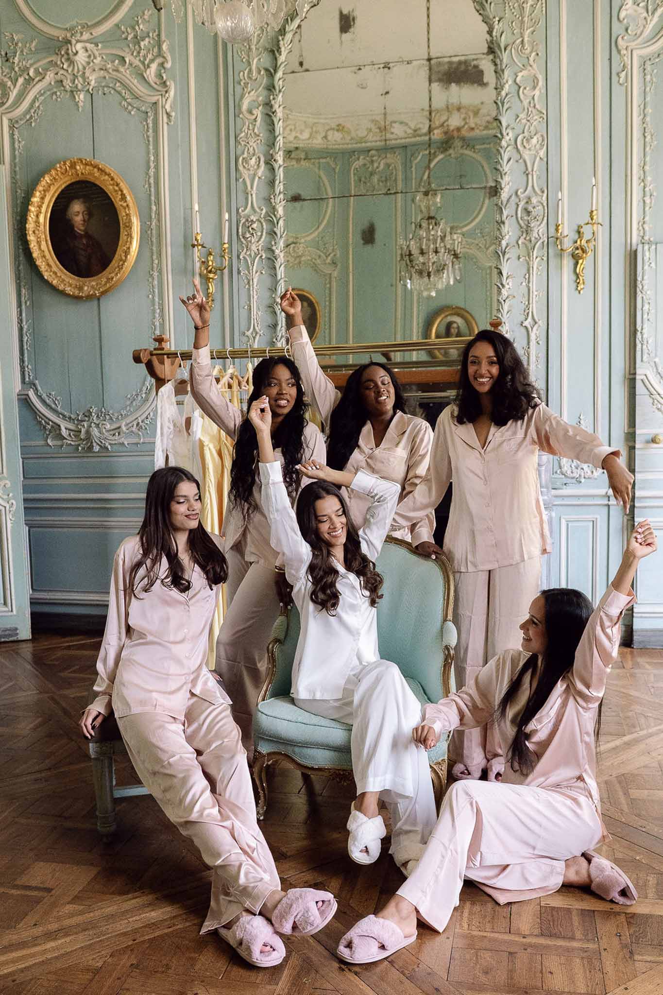 Bride and five bridesmaids in matching satin pajamas posing in ornate chateau room with blue paneling
