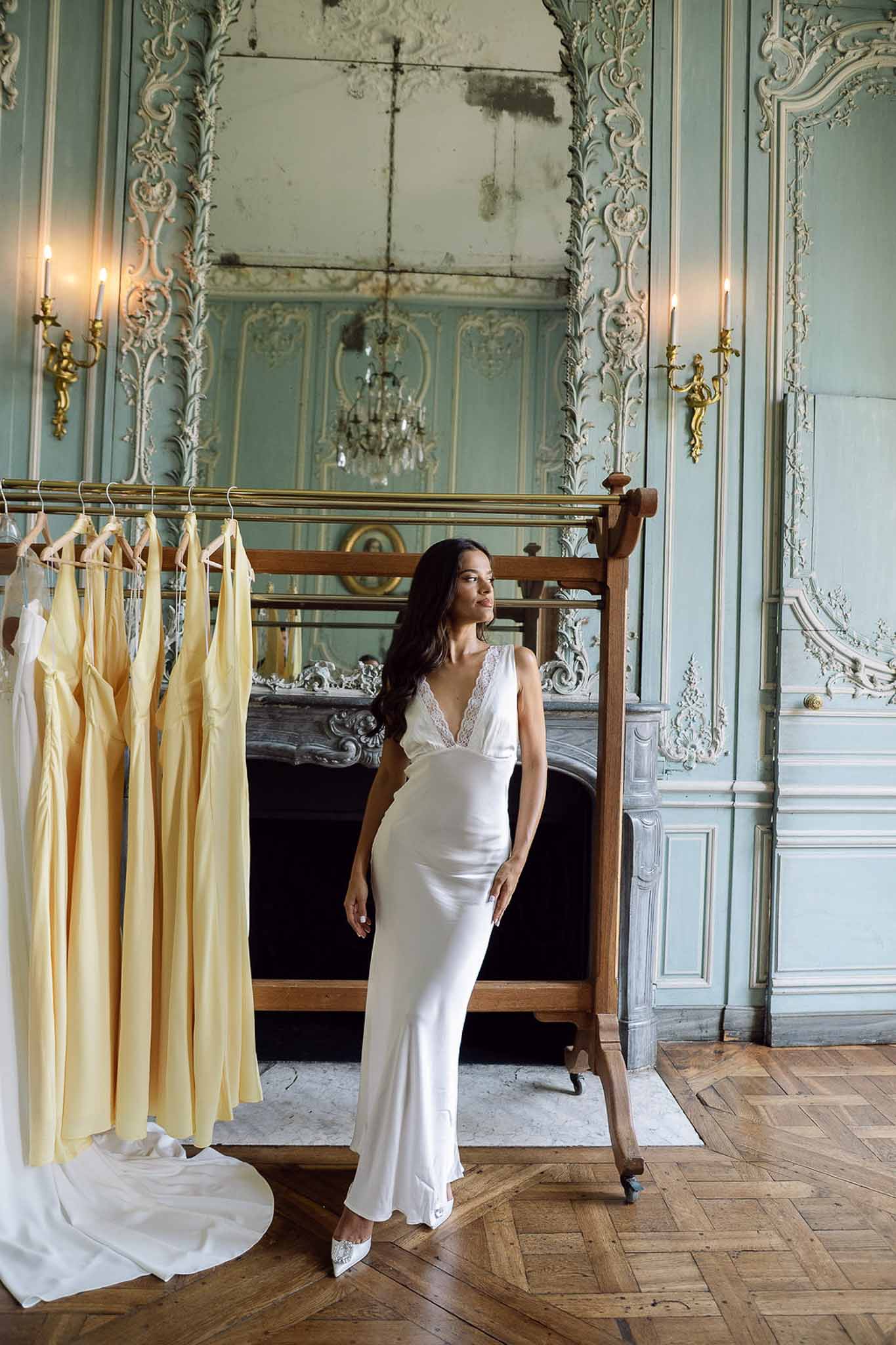 Bride in white satin slip dress beside antique mirror in chateau room with yellow bridesmaid dresses on rail