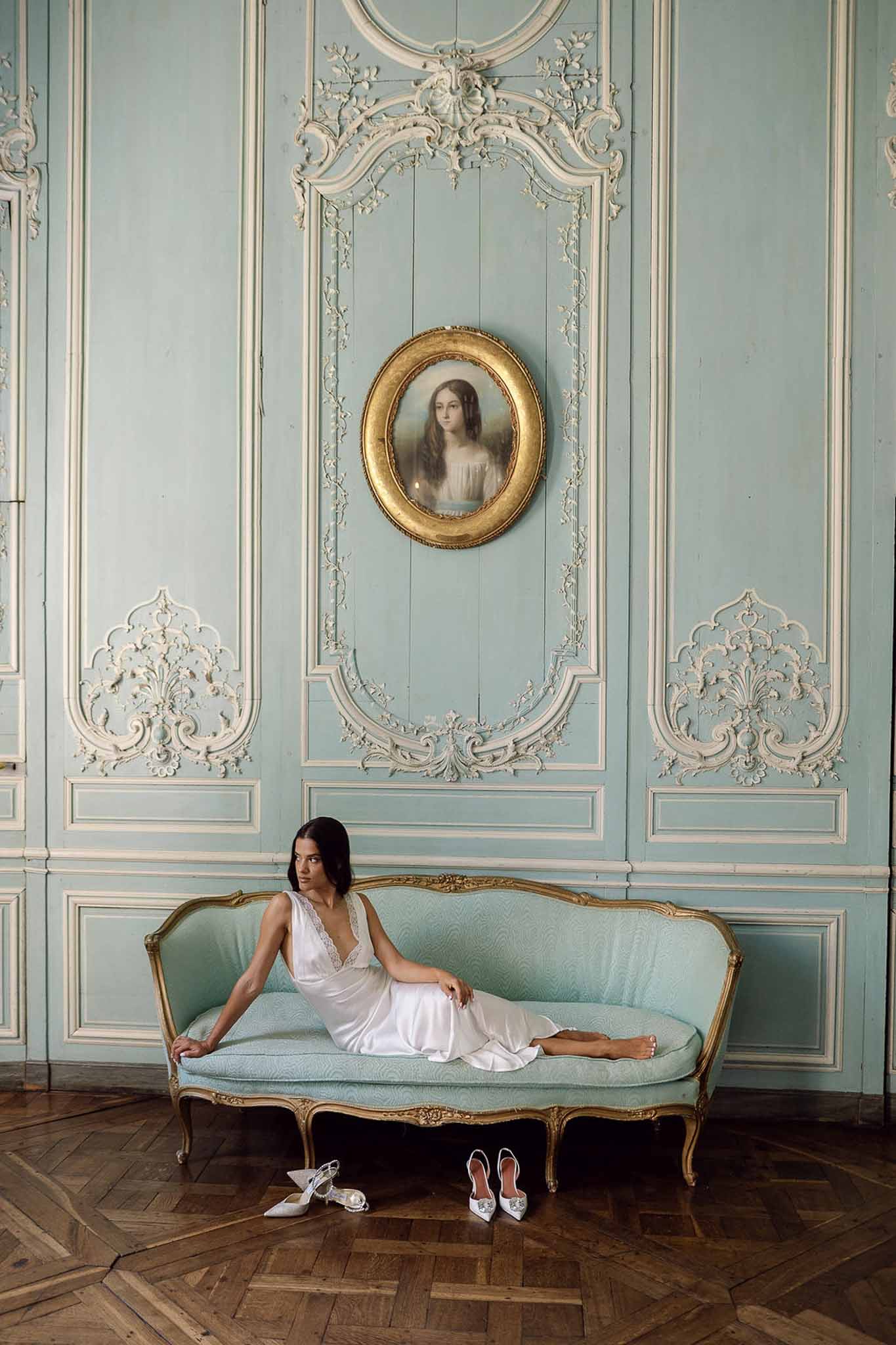 Bride in white satin slip dress on gilt canape beneath blue boiserie paneling and oval portrait painting