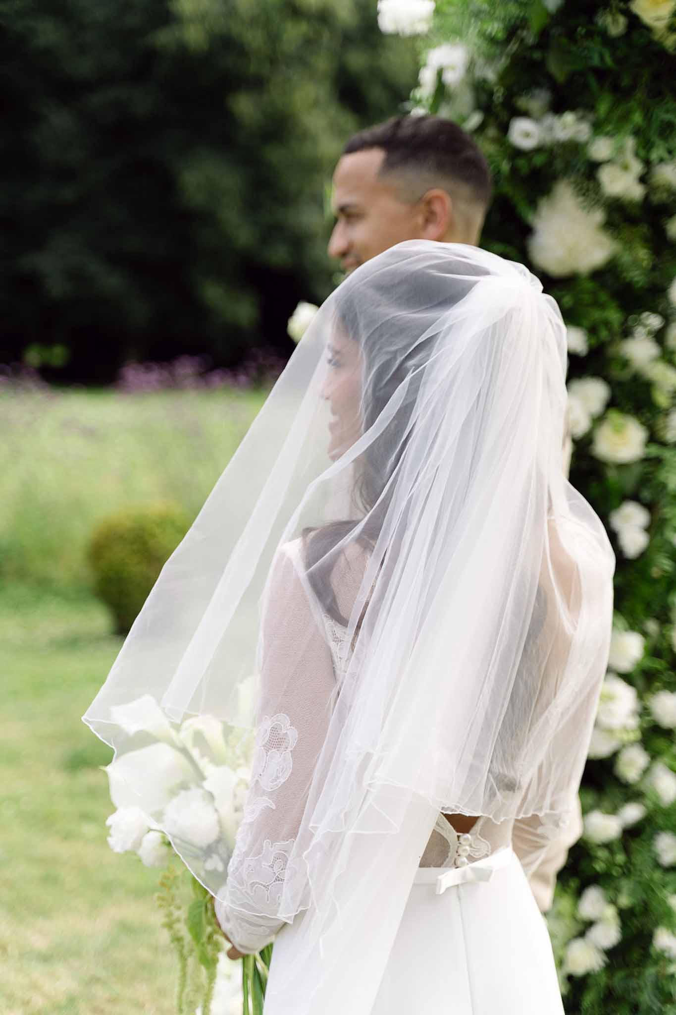 Bride and groom standing beside a white floral arch, veil draped over both, bride holding white tulip bouquet