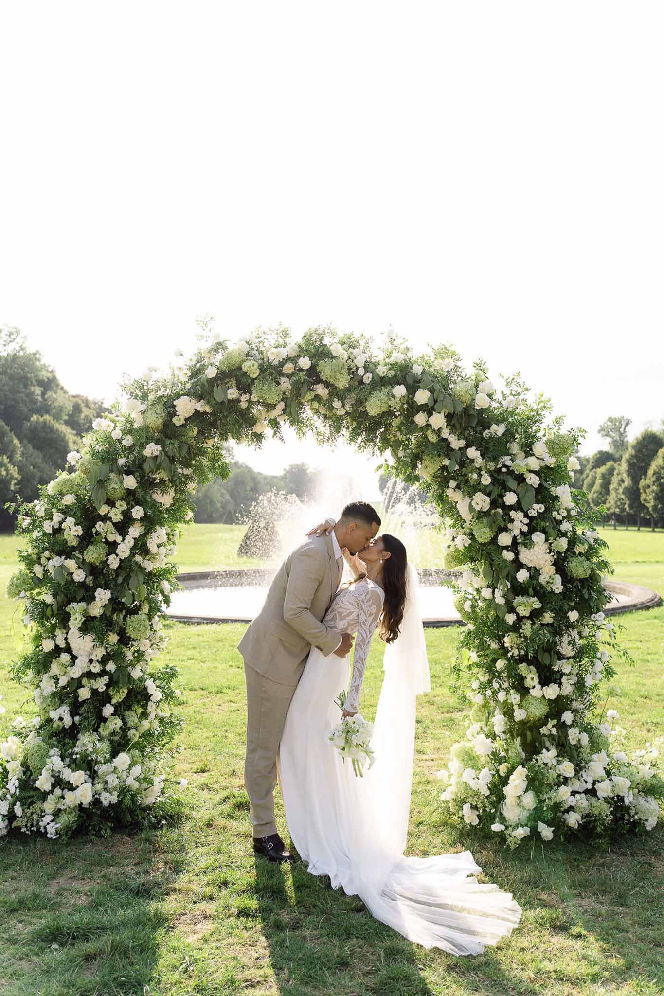Couple kissing beneath a circular arch of white roses and hydrangeas in a formal chateau garden