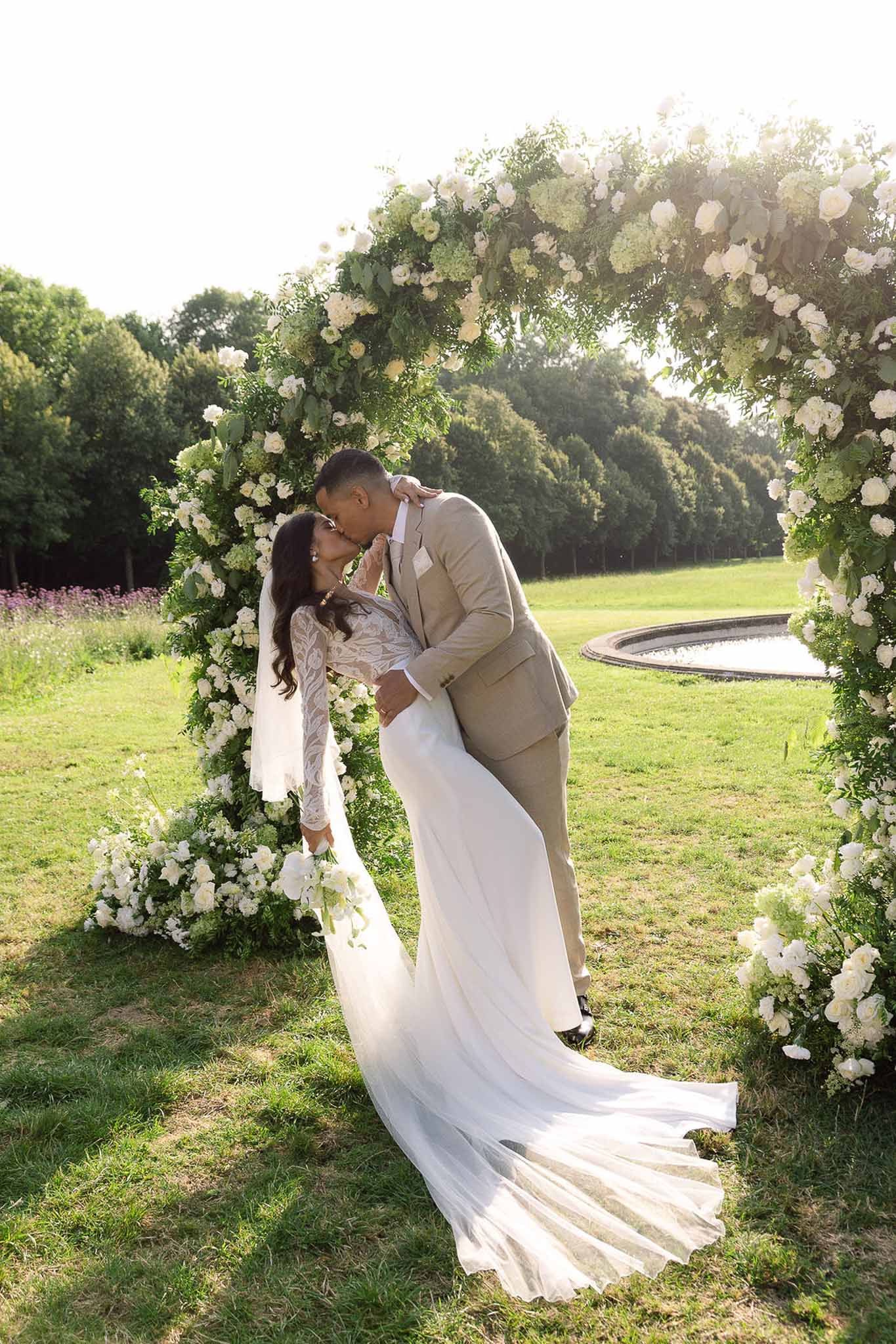 Couple shares dip kiss under circular white rose and hydrangea arch in chateau garden at golden hour