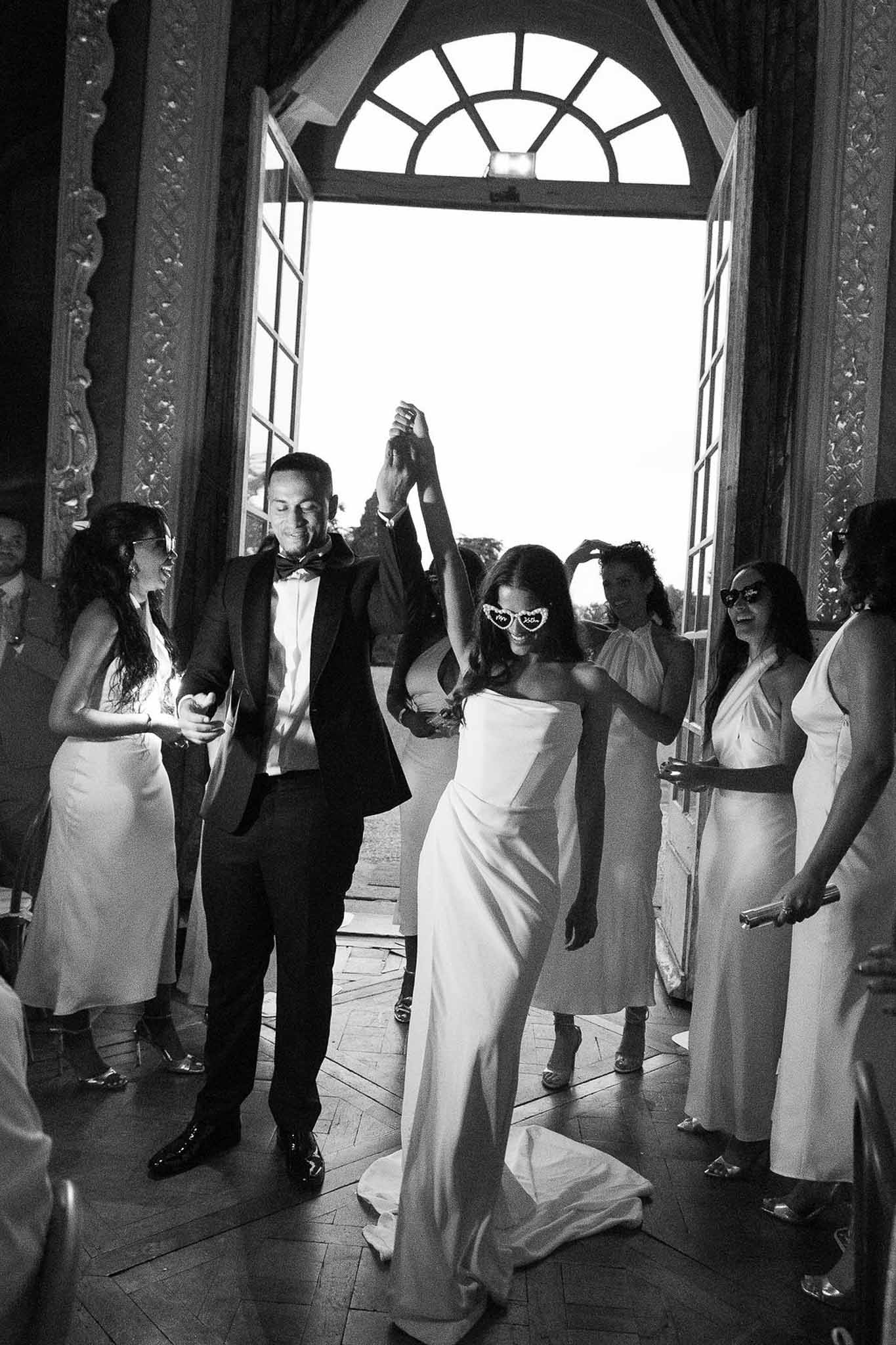 Black and white photo of couple entering chateau reception through French doors as guests cheer