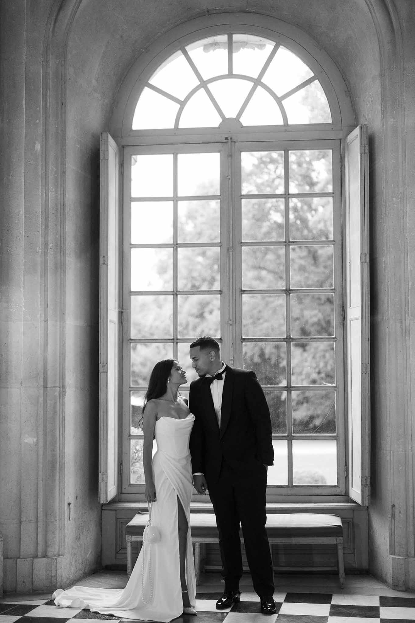 Black-and-white portrait of bride and groom standing close on checkered floor before a tall arched chateau window