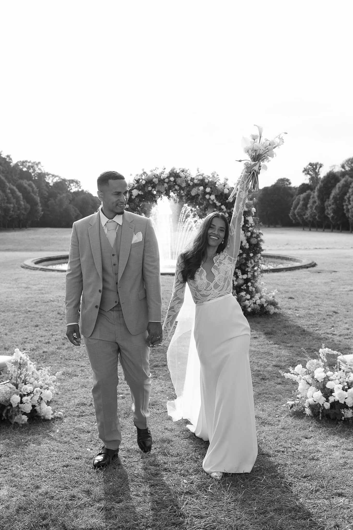 Black-and-white portrait of bride and groom walking across lawn after ceremony, bride laughing with bouquet raised