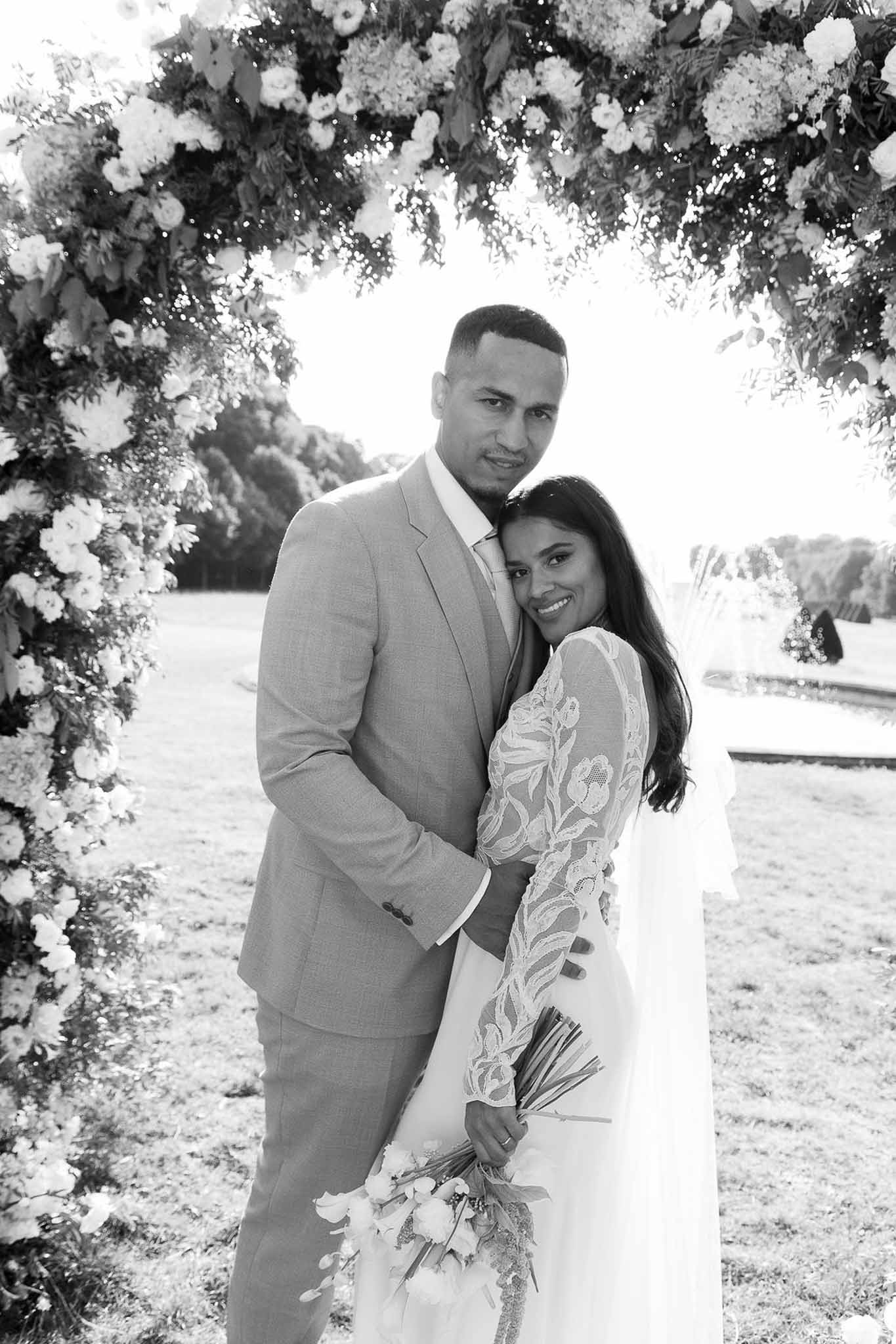 Black and white of couple under dense floral arch, bride in long-sleeve lace gown with orchid bouquet in formal garden
