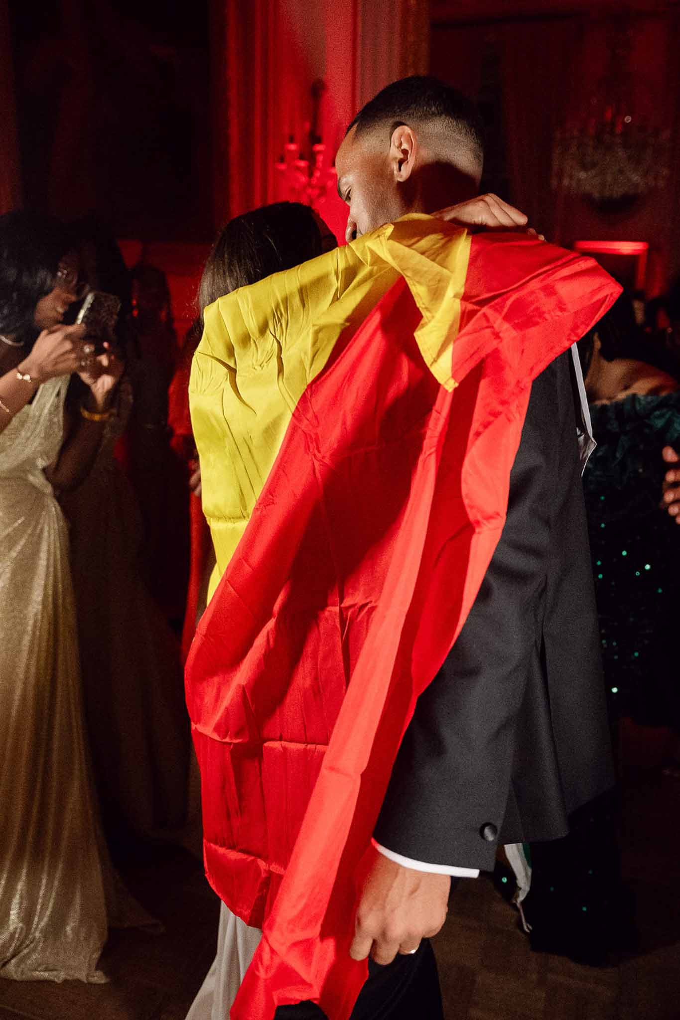 Couple embraces on dance floor wrapped in flag under red ambient lighting in chateau ballroom
