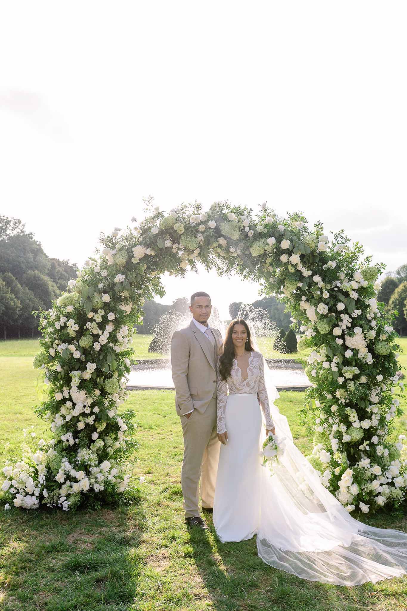 Couple beneath circular white rose and hydrangea arch with garden fountain in background