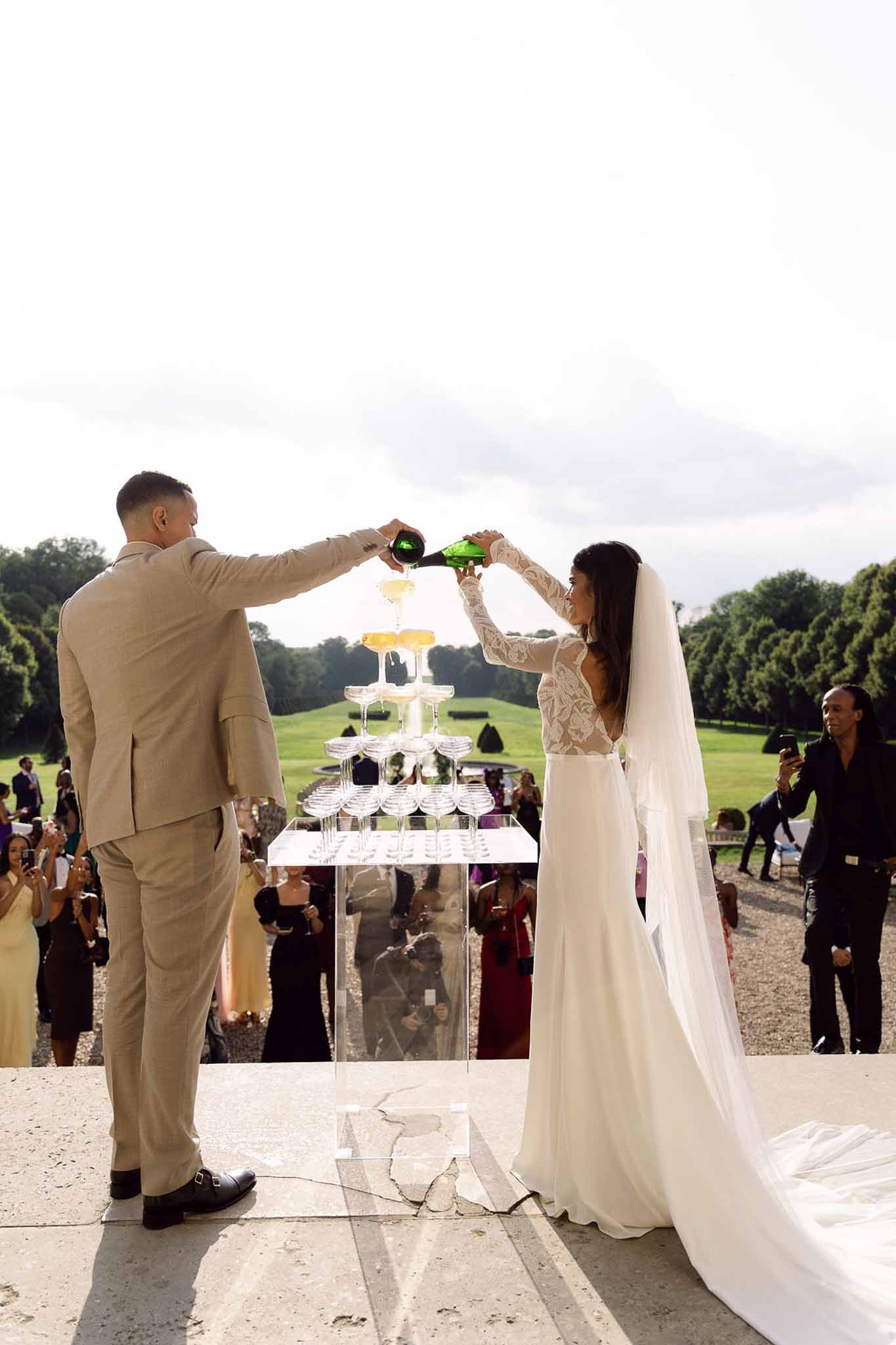 Couple pouring champagne tower on acrylic pedestal on chateau terrace with 30 guests below