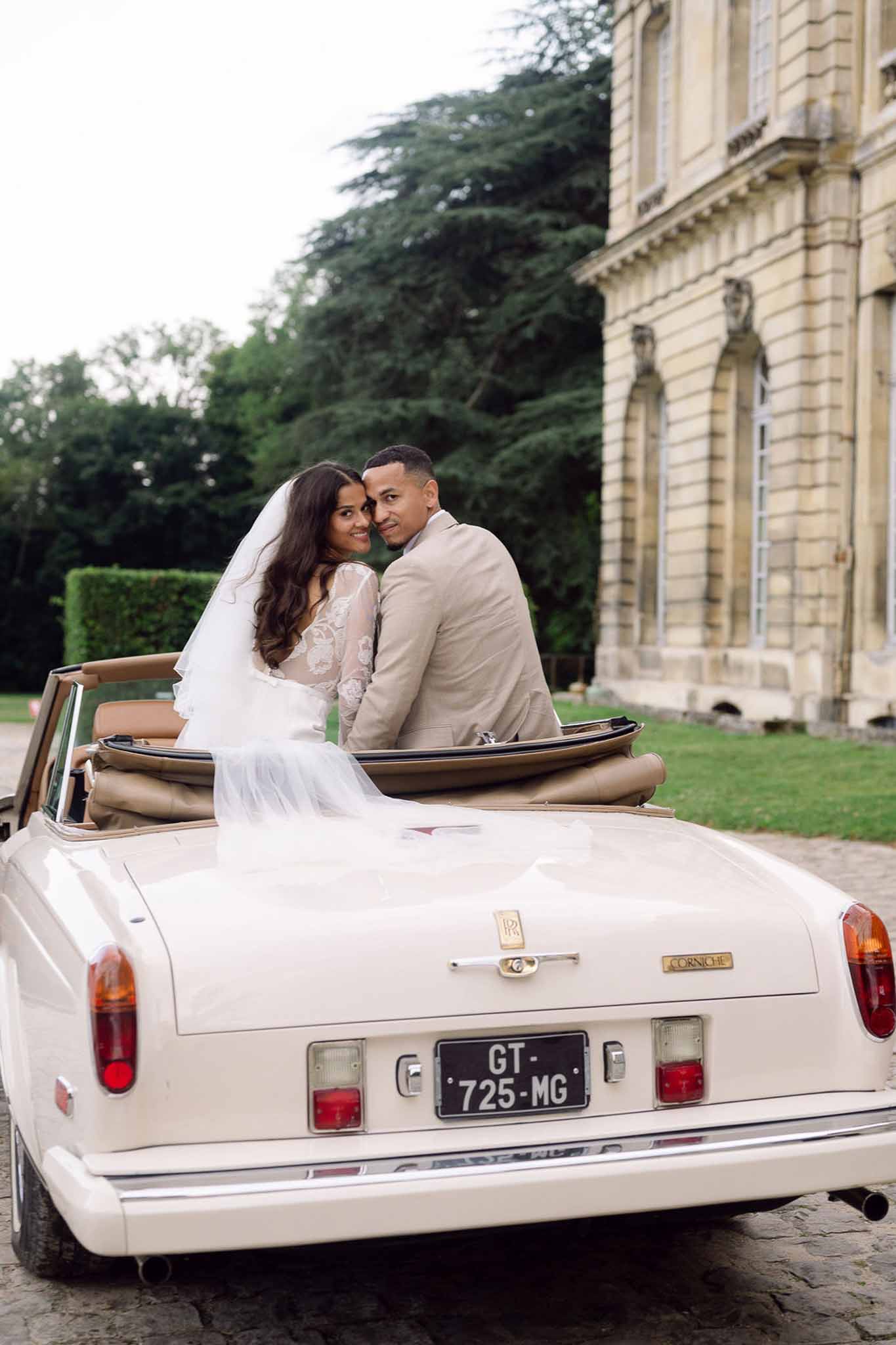 Bride and groom seated in cream convertible Rolls-Royce looking back at camera with chateau facade behind
