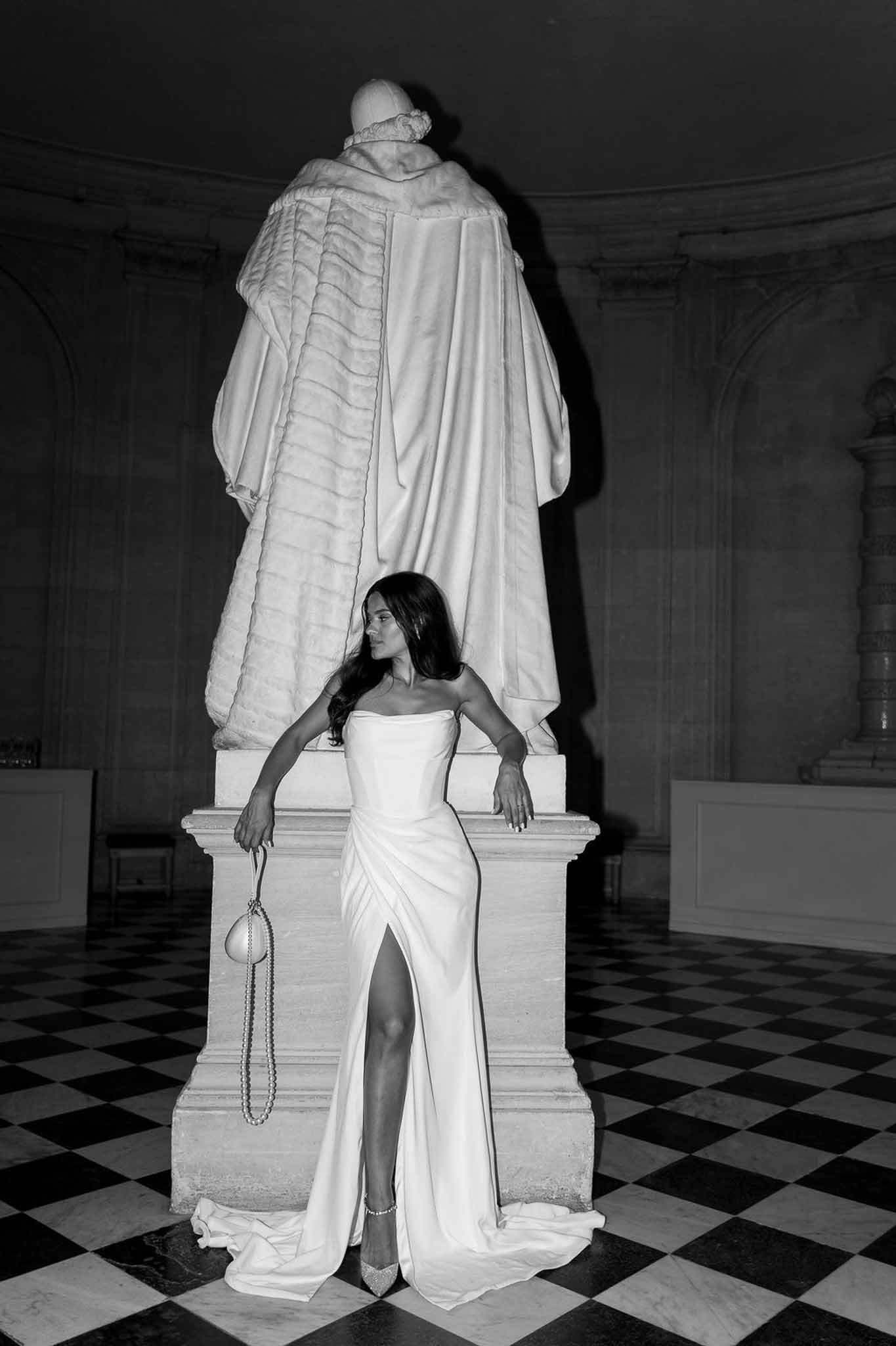 Bride in strapless satin gown posing beside marble statue on checkered floor in black and white