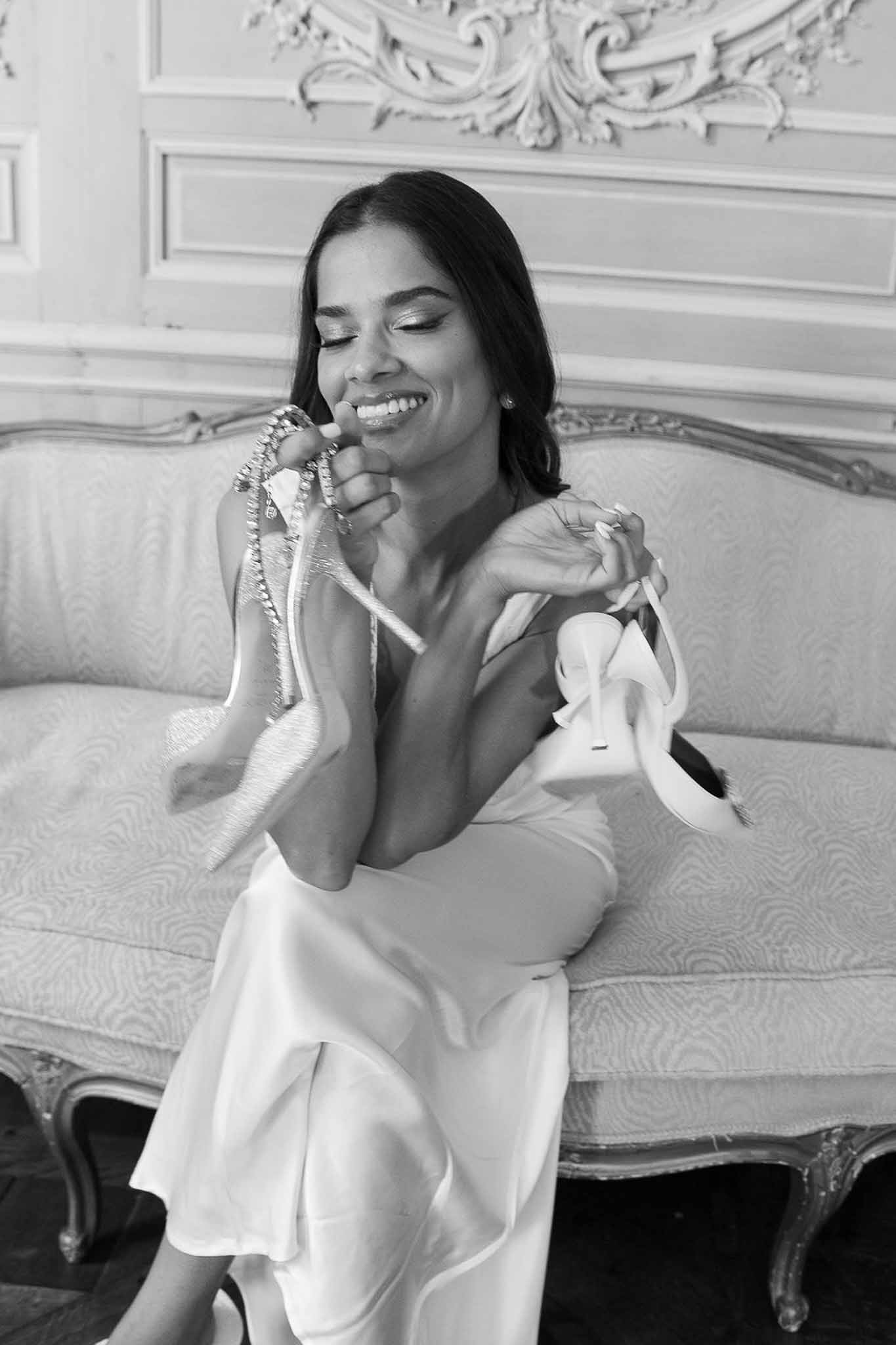 Black-and-white portrait of bride laughing on ornate sofa holding crystal-embellished heels in a French chateau interior