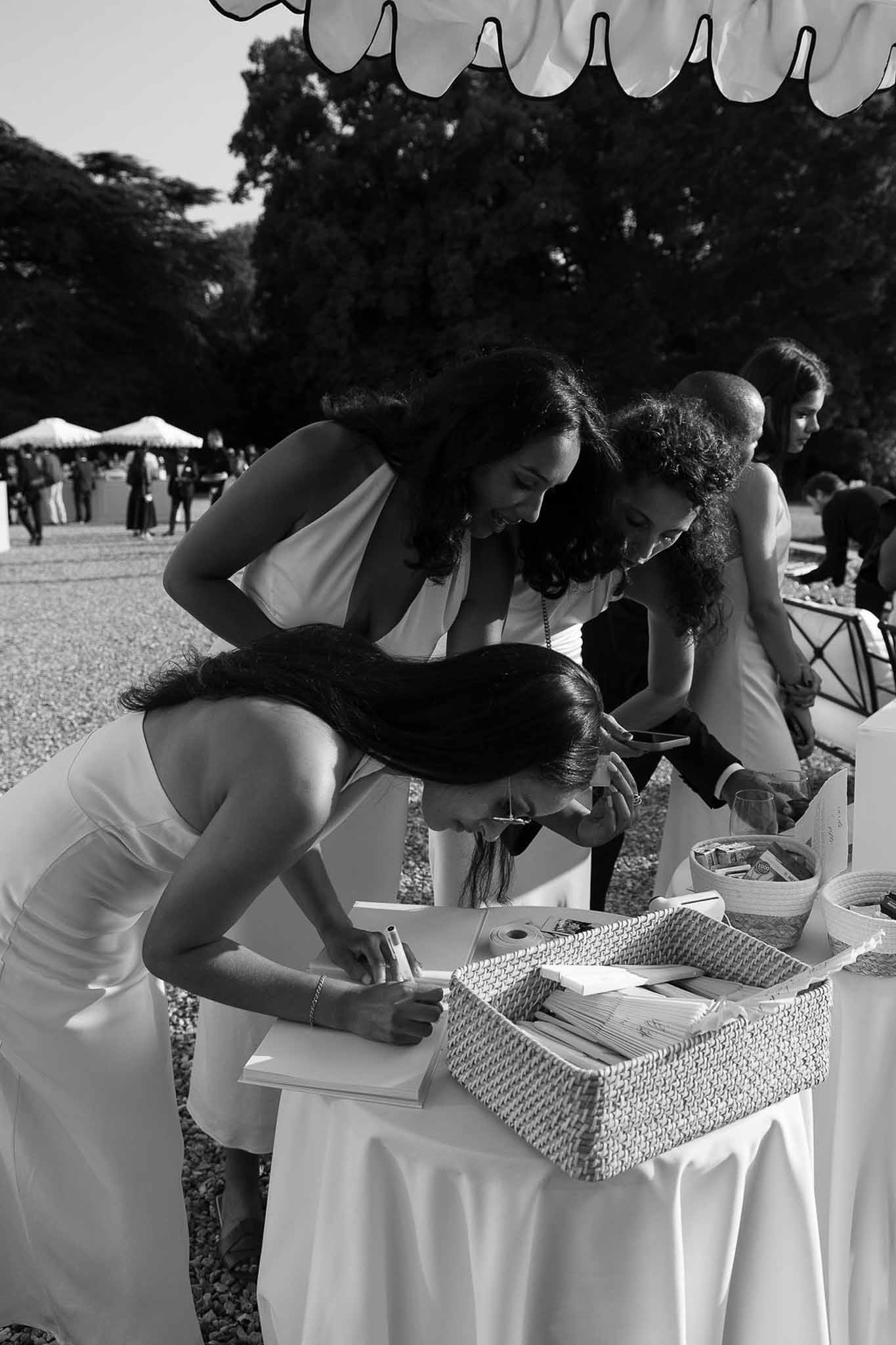 Black and white candid of three women signing guestbook at outdoor cocktail station under canopy