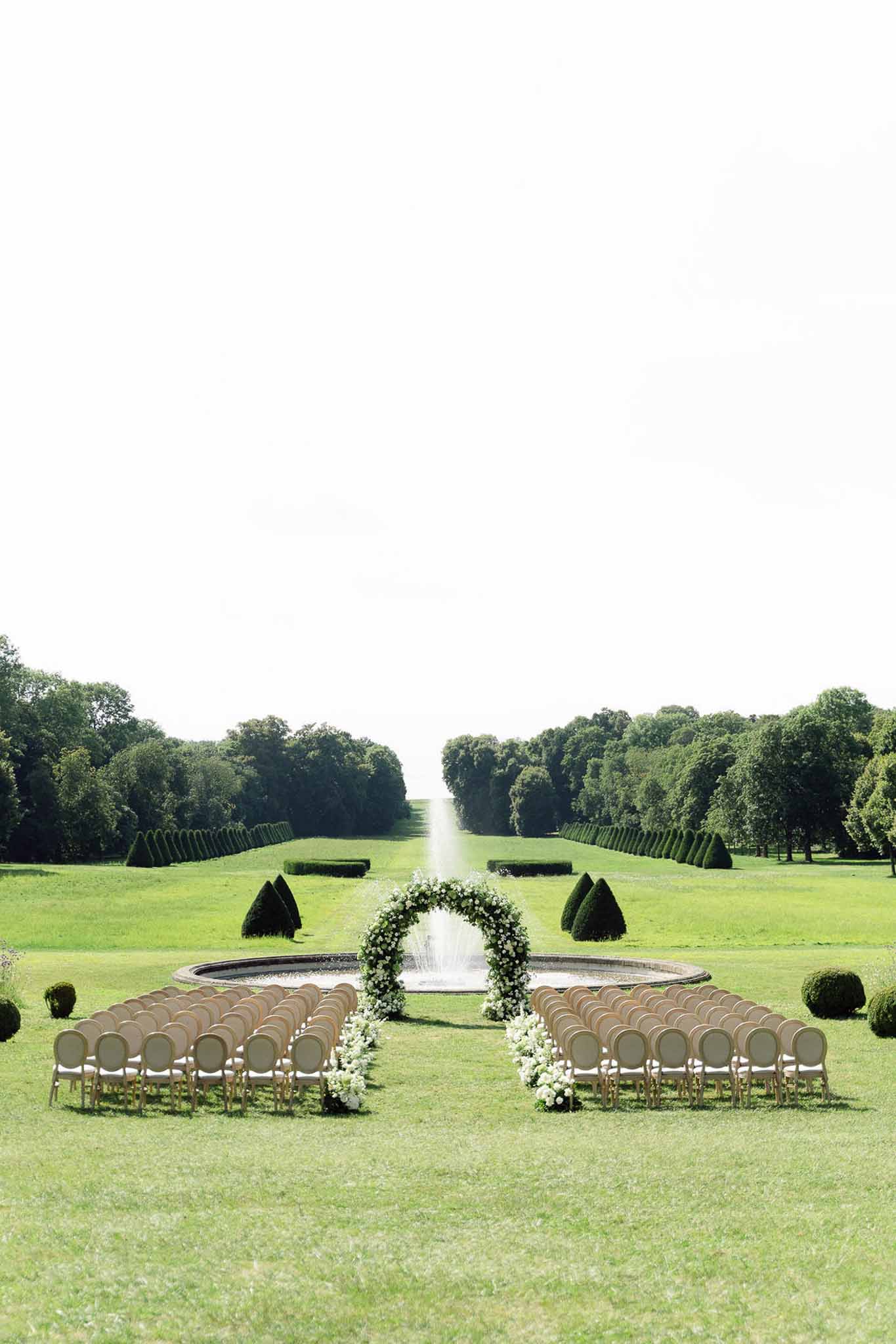 Outdoor ceremony setup with gold chairs along a formal garden allee and circular white floral arch framing a fountain