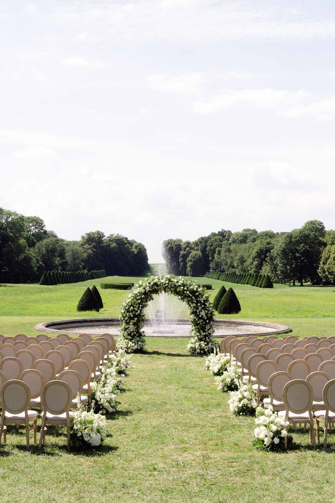 Outdoor ceremony setup with gold chairs, white floral arch over fountain, and formal French estate garden