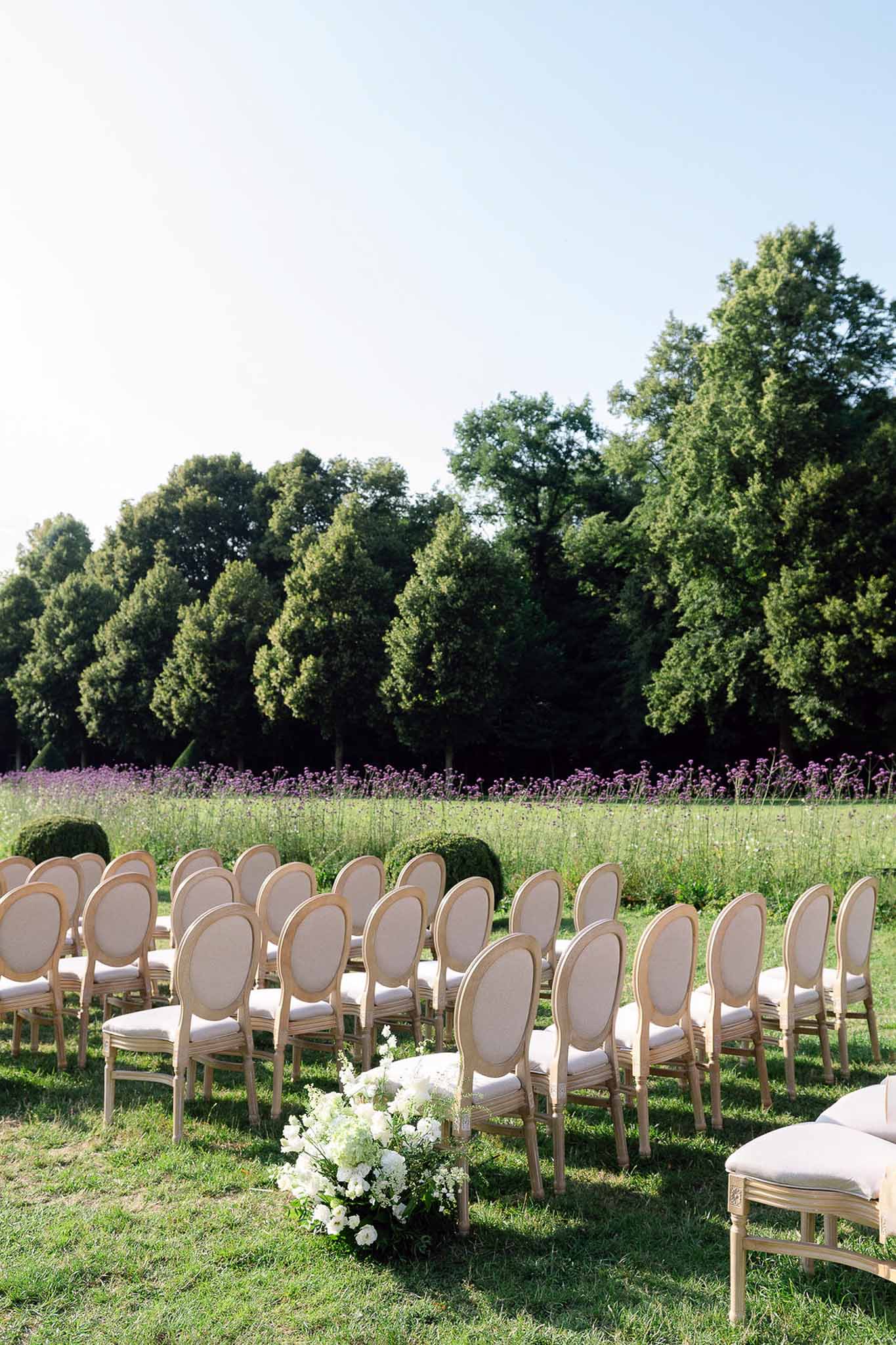 Outdoor ceremony setup with cream upholstered Louis XVI chairs on lawn and white hydrangea aisle markers