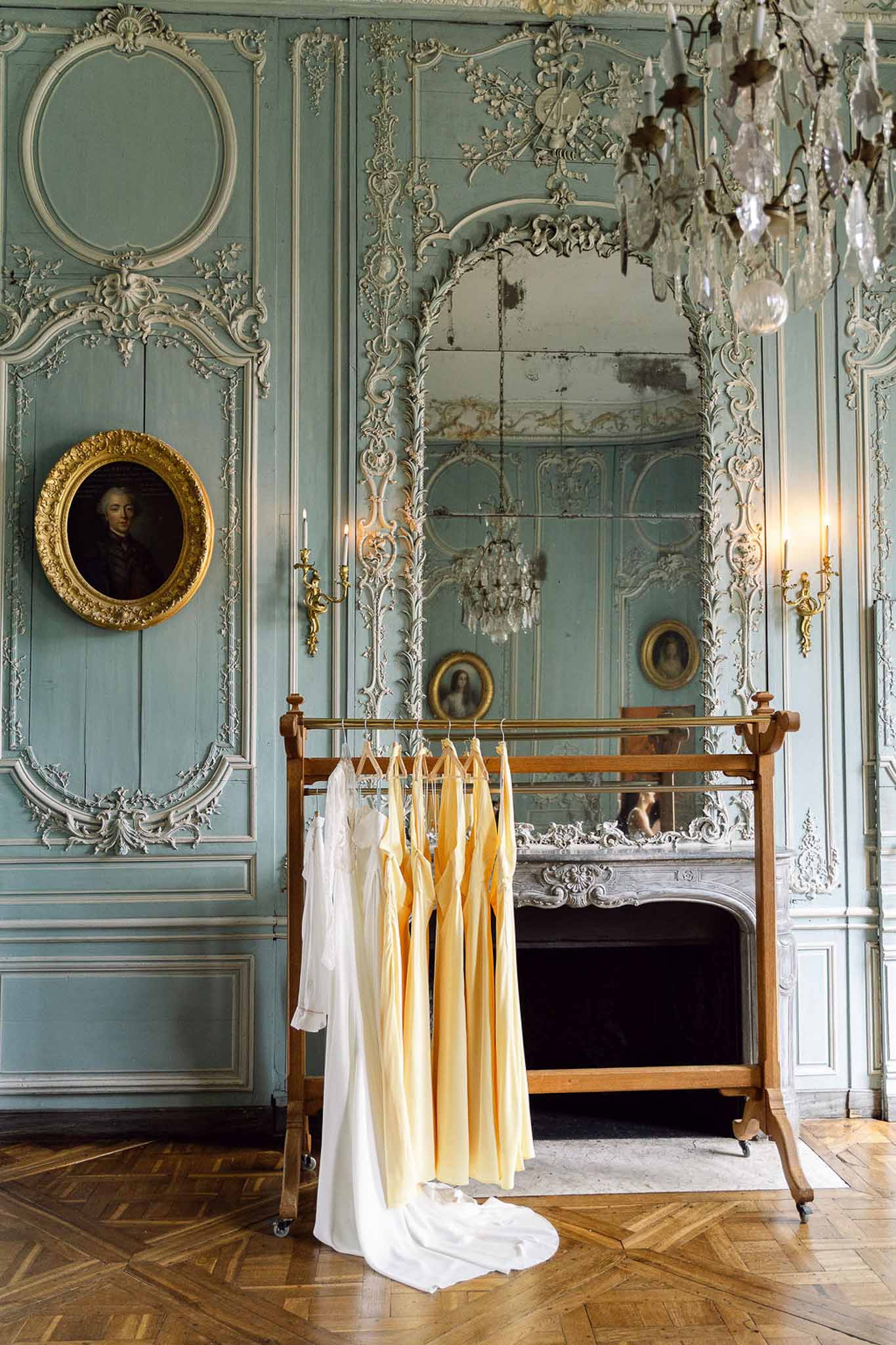 Bridal gown and four yellow bridesmaid dresses on garment rack before blue boiserie fireplace and mirror