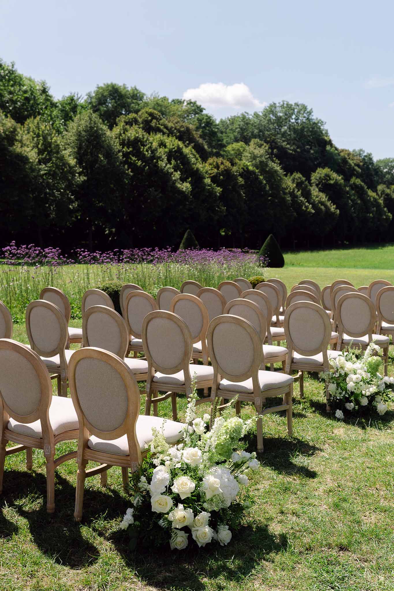 Louis-style chairs with white rose aisle markers on lawn with lavender border and topiary in background