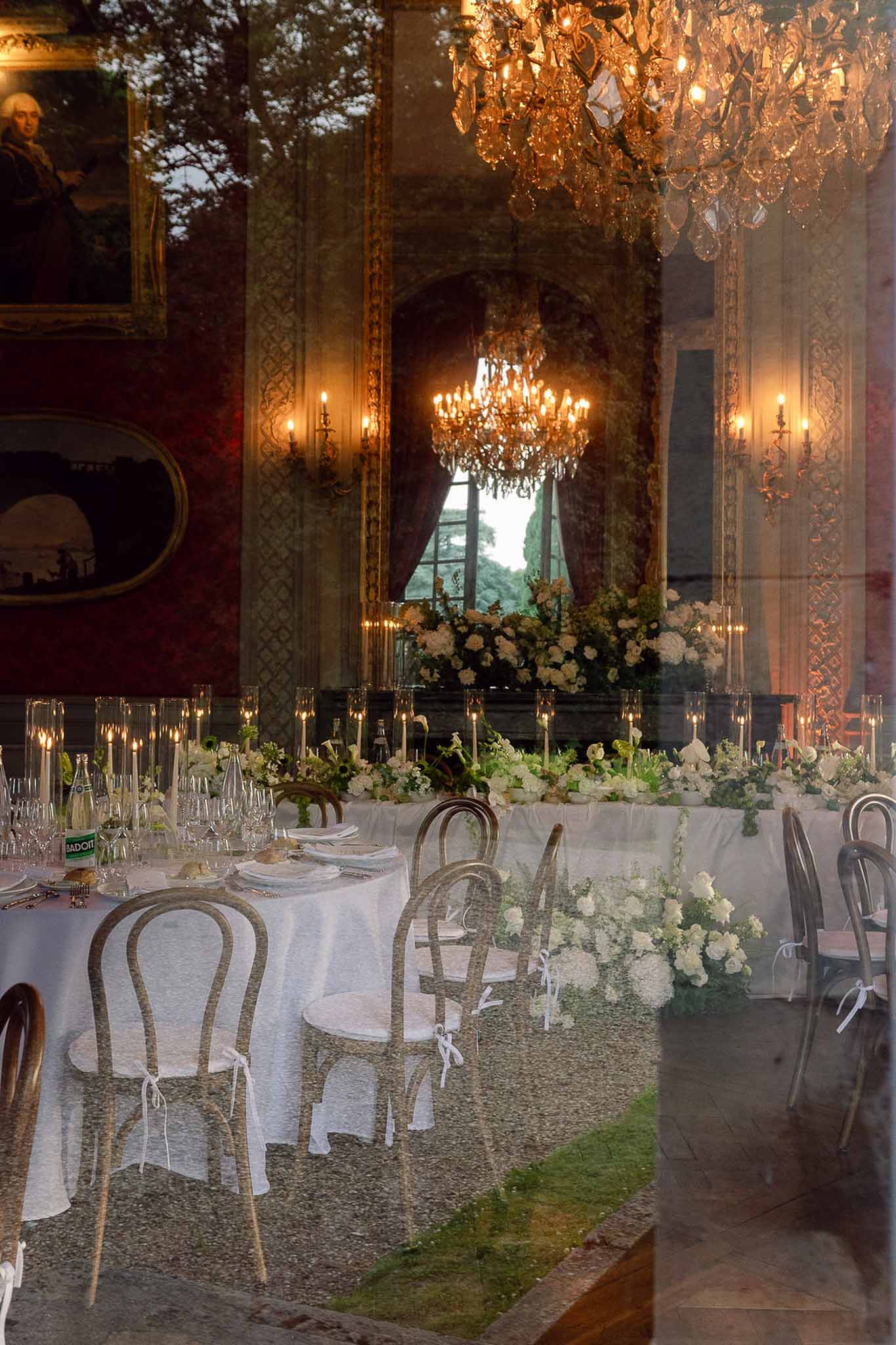 Chateau ballroom reception through glass with chandeliers, white hydrangea centrepieces, and taper candles