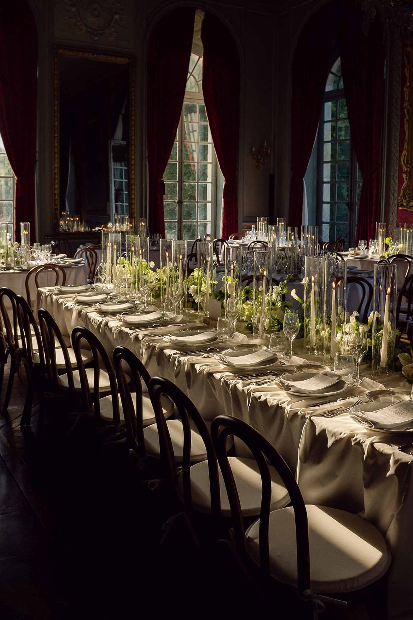 Long banquet table with white calla lily runner and hurricane candles in gilded chateau ballroom with crimson drapes