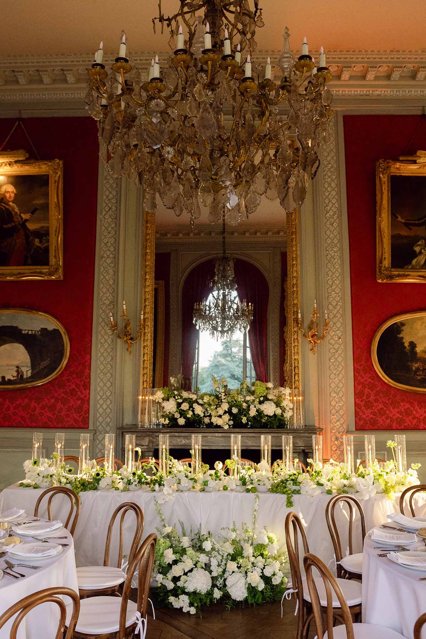 Wedding reception head table with white hydrangea garland inside a grand red-walled room with crystal chandelier