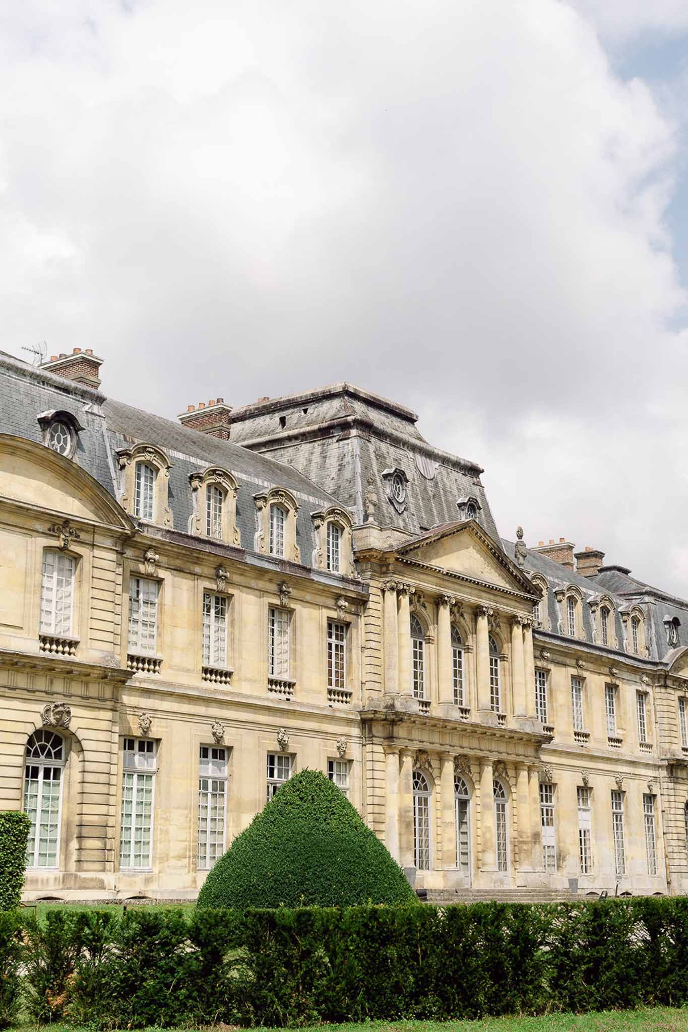 Cream limestone classical chateau with arched windows, carved pediment, and conical topiary in parterre
