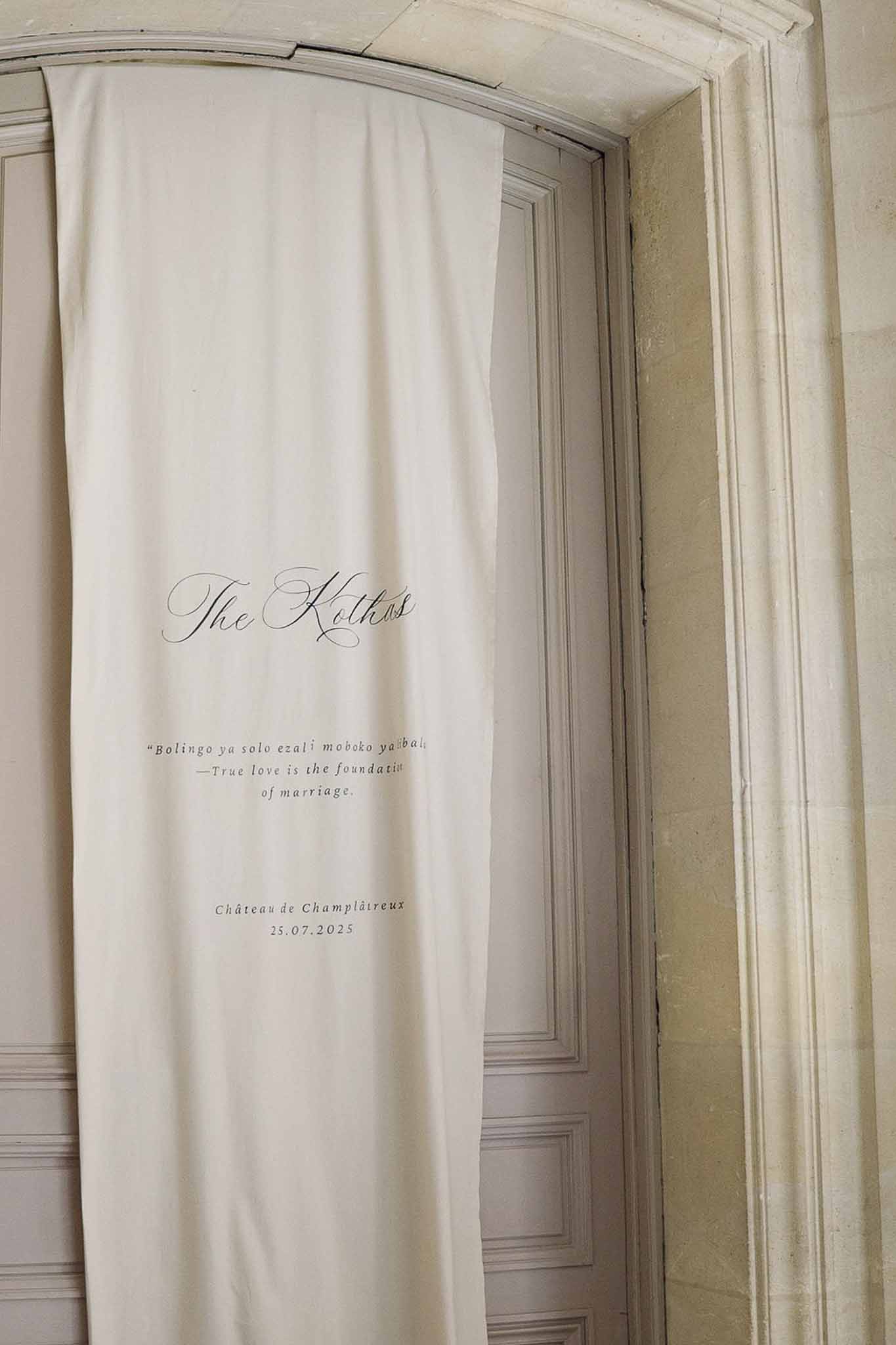 Ivory fabric wedding banner with calligraphy names and bilingual Lingala-English quote hanging in stone chateau doorway