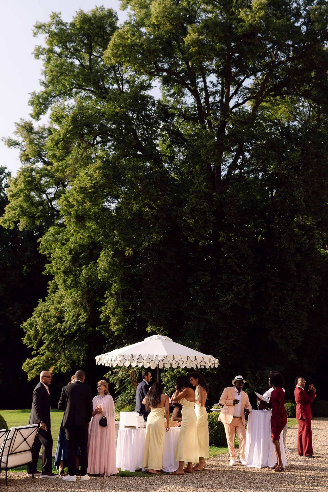 Guests mingling on a gravel terrace during cocktail hour under a white market umbrella in afternoon sun
