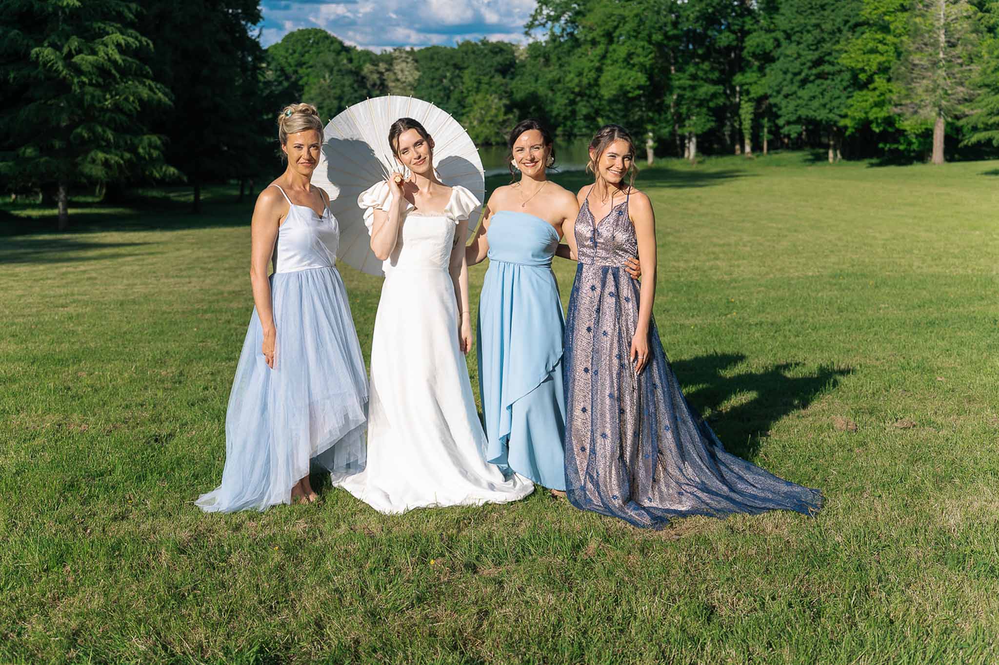 Bride with white parasol and three bridesmaids in mismatched blue dresses on park lawn by lake