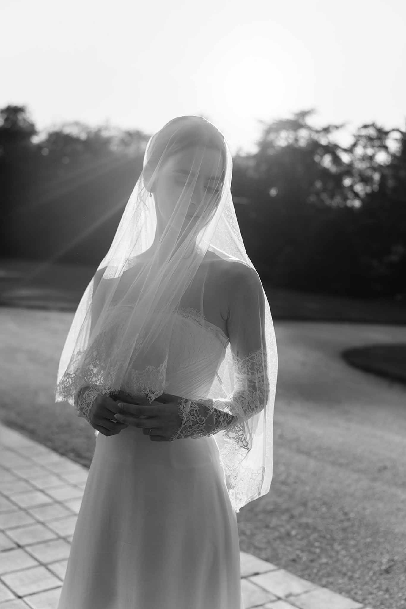 Backlit bride with lace-bordered veil draped over head and strapless gown on terrace in B&W
