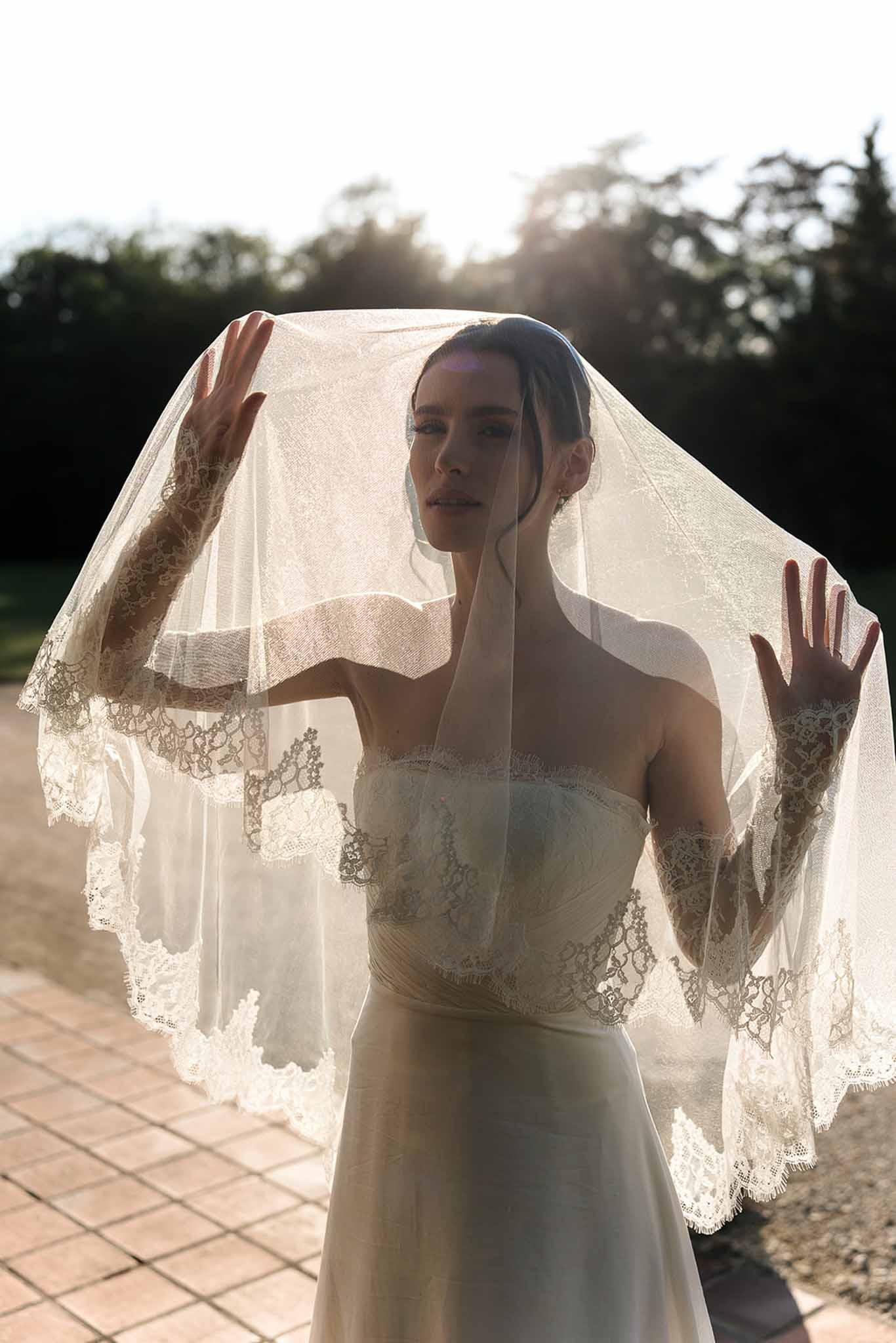 Bride holding lace-trimmed cathedral veil above her head in backlit sunset portrait on terrace