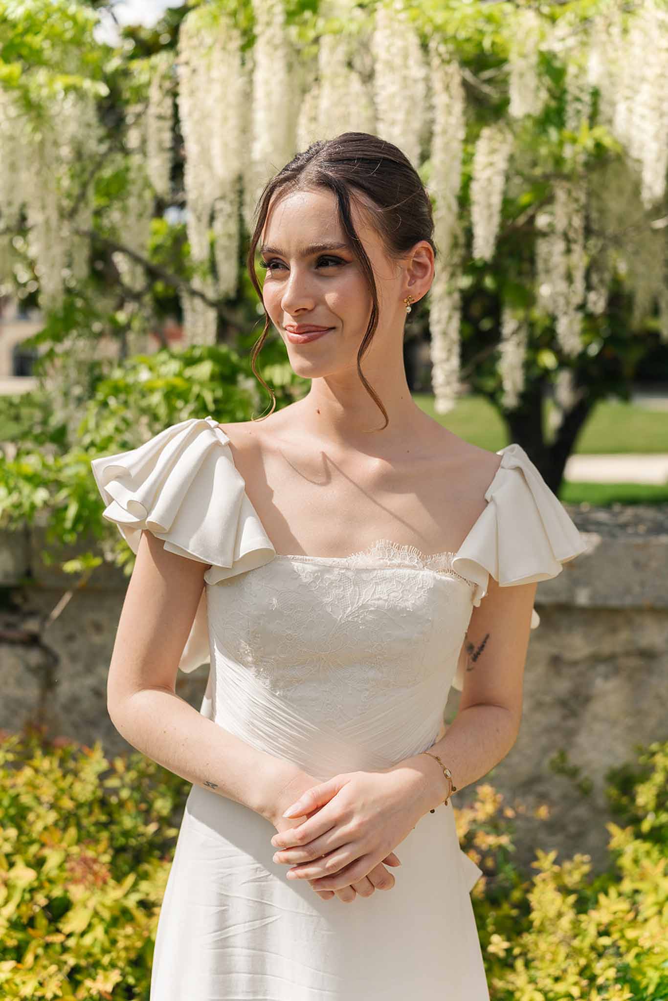 Bride in ivory ruffle-sleeve lace gown smiling beneath white wisteria tree in garden setting