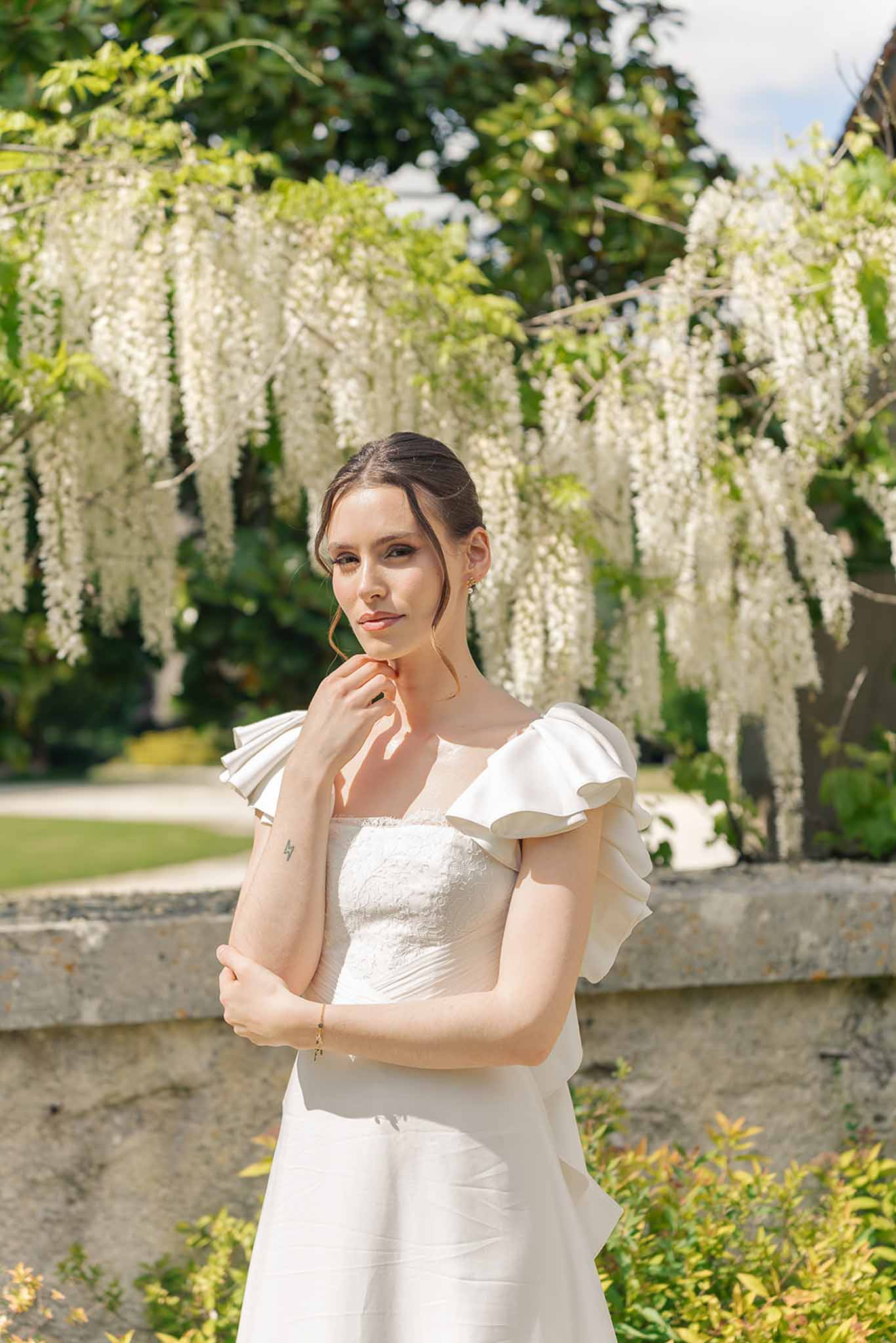 Bride in off-shoulder ruffle-sleeve gown with low updo before white wisteria and stone balustrade