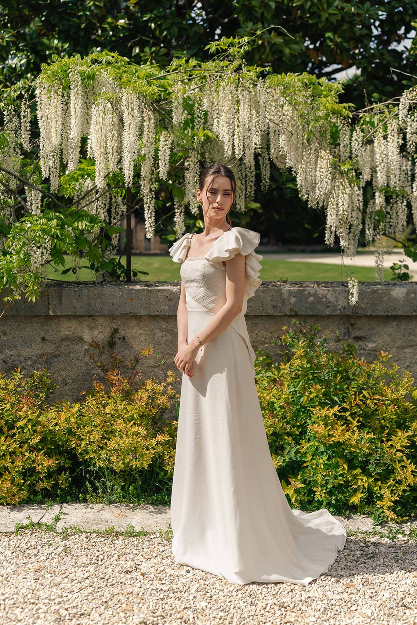 Bride in ivory A-line gown with ruffled cap sleeve standing before wisteria-draped stone balustrade in chateau garden