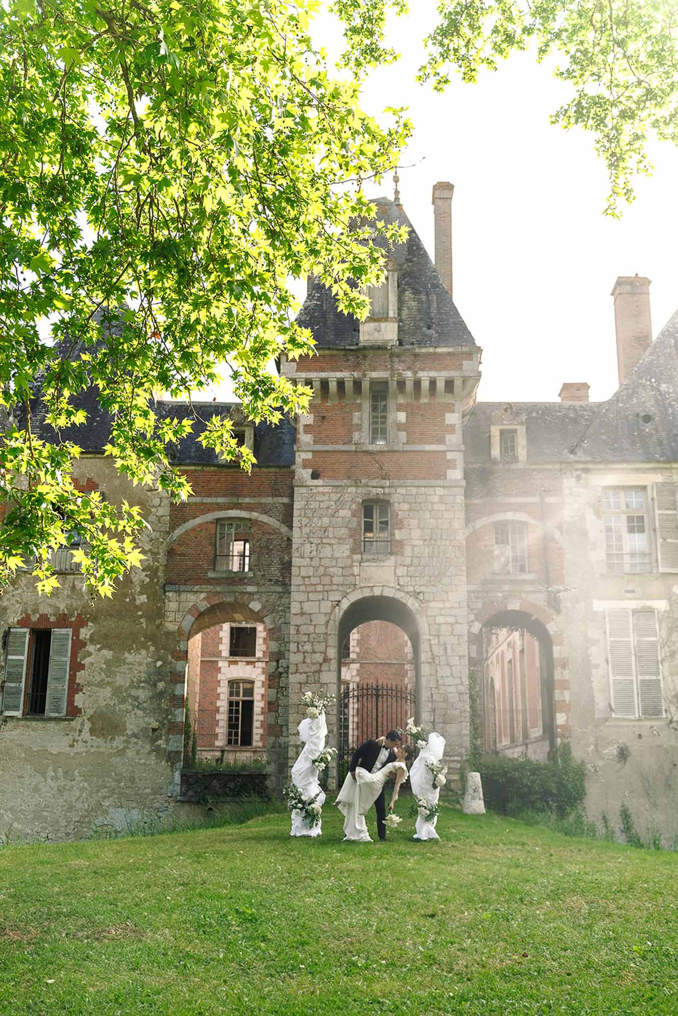 Groom dips bride for kiss at chateau archway flanked by attendants styled as classical statues with white florals