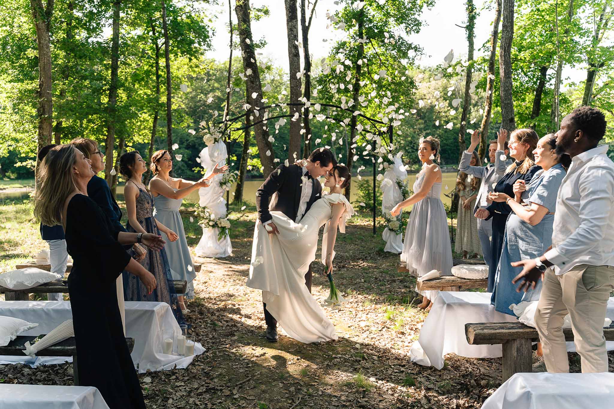 Bride and groom dip kiss walking down aisle as guests toss white petals in woodland ceremony clearing
