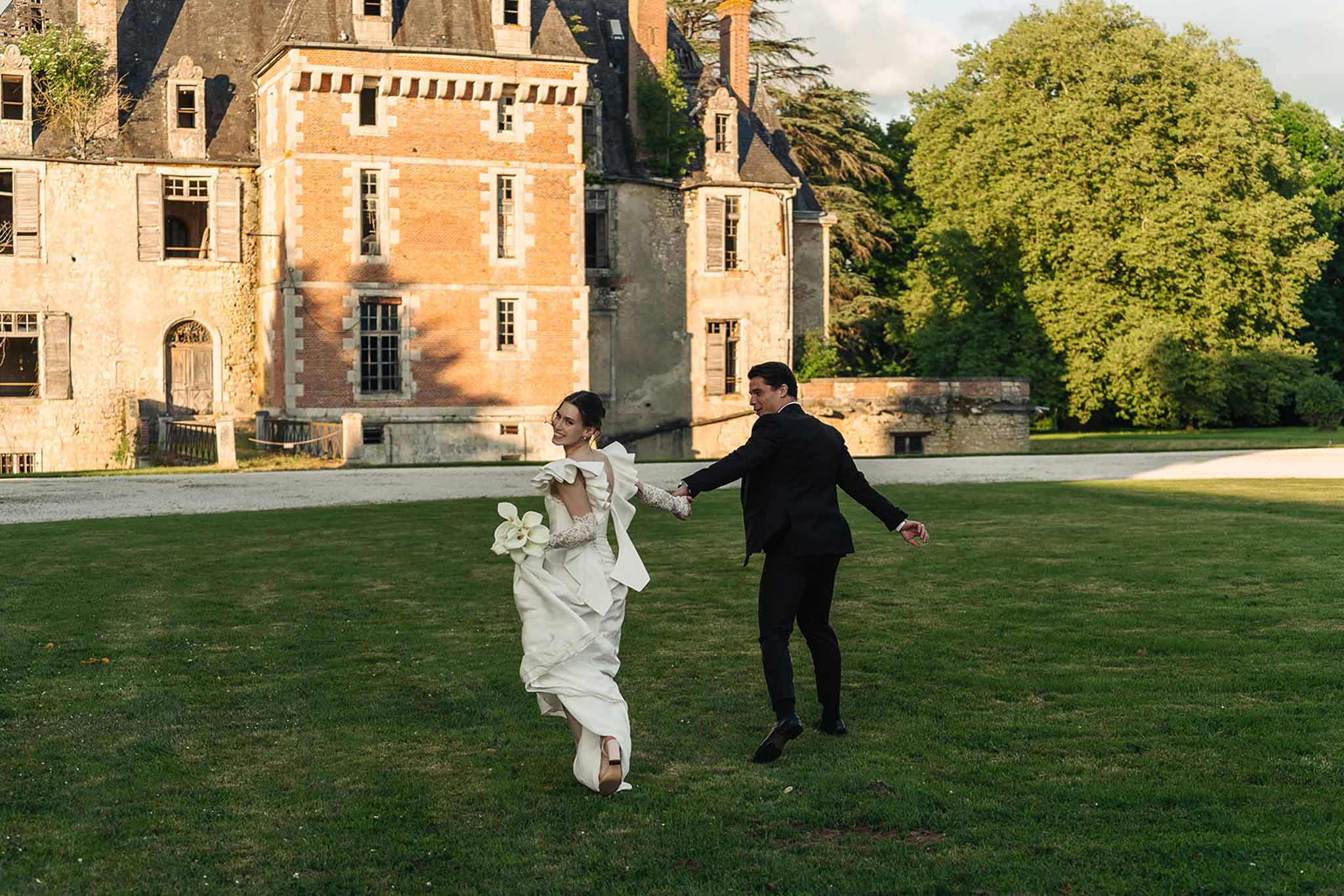 Couple runs across lawn in golden light with bride's sculptural bow gown and calla lily bouquet at chateau
