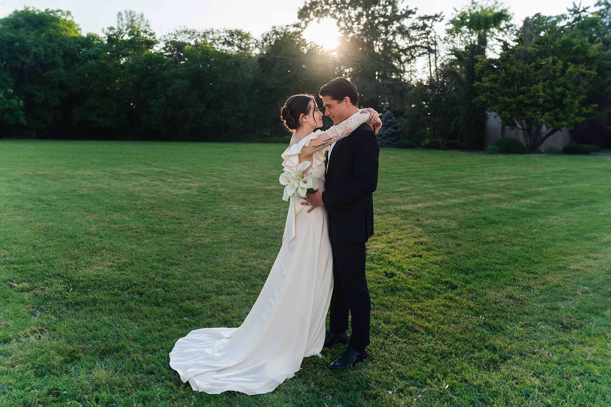 Bride in ruffled white gown and groom in black tuxedo touching foreheads on open lawn at golden hour