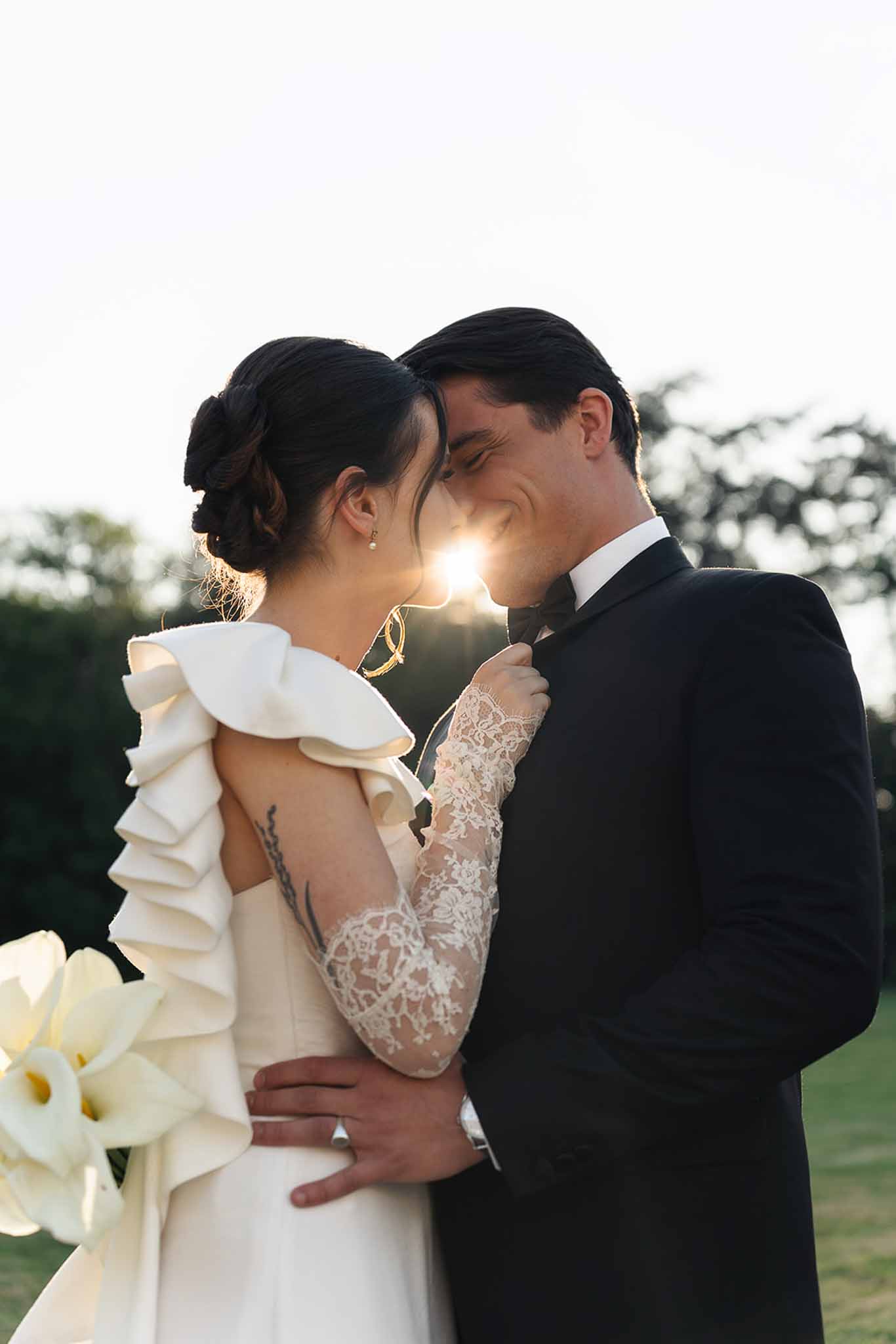 Bride in tiered ruffle gown with lace sleeves and groom in tuxedo touch foreheads in golden backlight