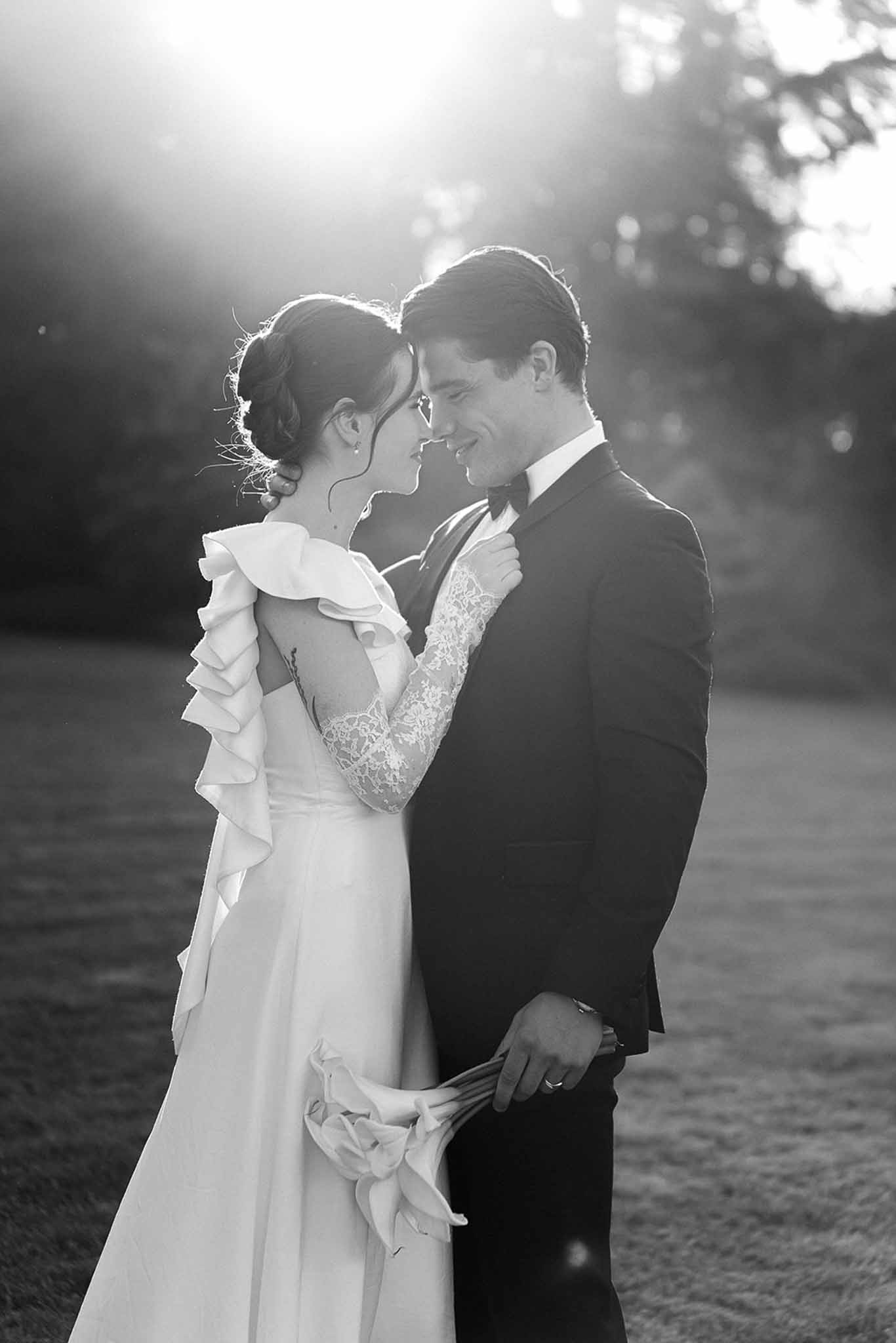 Black and white backlit portrait of couple with foreheads touching, bride holding calla lily bouquet at golden hour